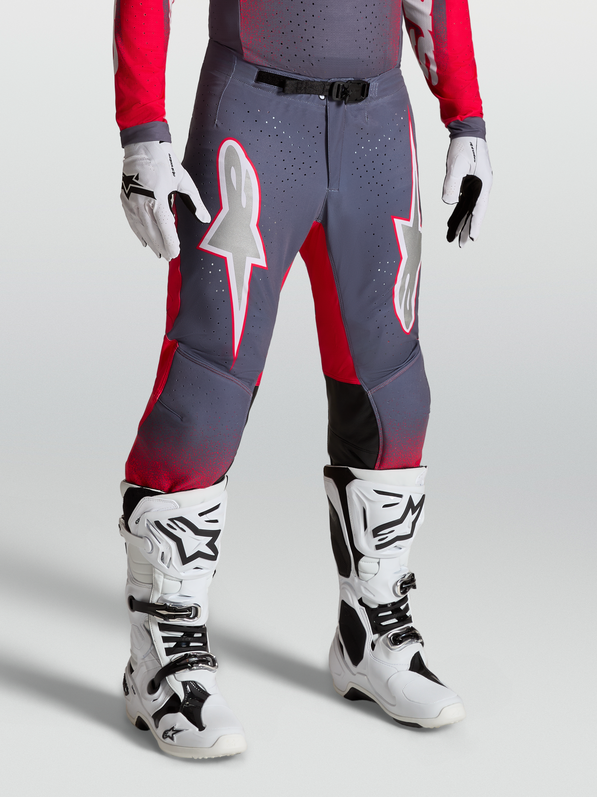 Alpinestars Pantaloni Supertech Hiten AFD, pantaloni da motocross grigi e rossi con grafiche a stella e fibbia magnetica, indossati con stivali bianchi.