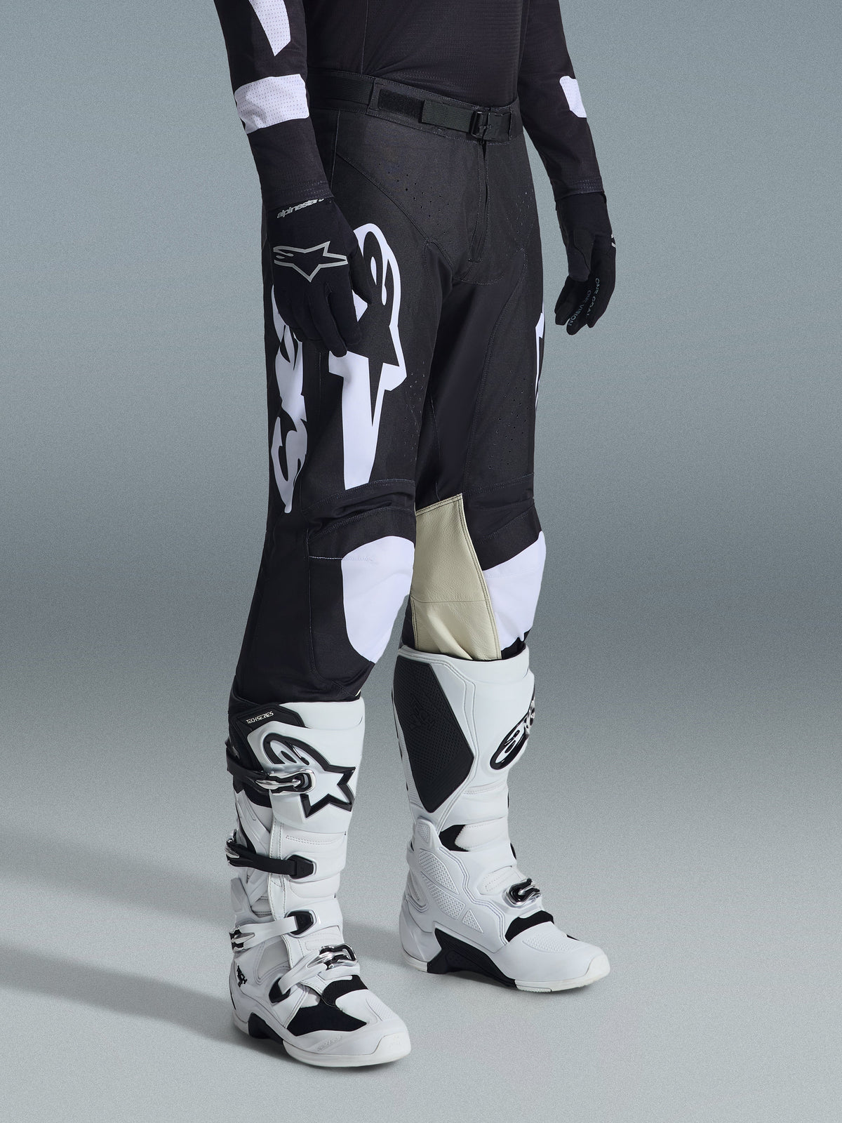 Pantaloni Alpinestars Racer Air Riway 2026, pantaloni da motocross neri e bianchi con grandi grafiche del logo bianco, pannelli interni delle ginocchia in pelle beige, indossati con stivali off-road bianchi e neri e guanti neri in studio.