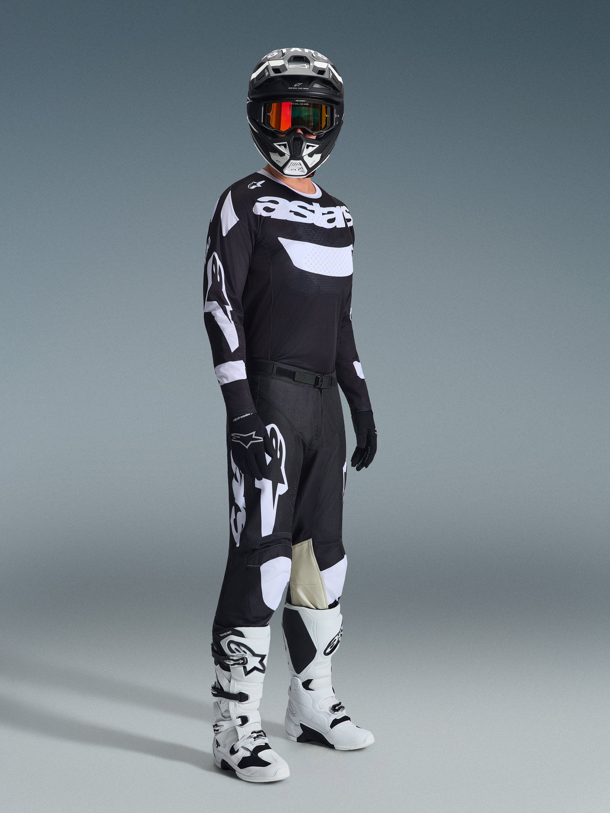 Pilota di motocross che indossa i pantaloni 2026 Racer Air Riway, nero e bianco, maglia coordinata con logo Alpinestars, casco nero con occhiali, stivali bianchi e neri, set completo per prestazioni motocross off-road