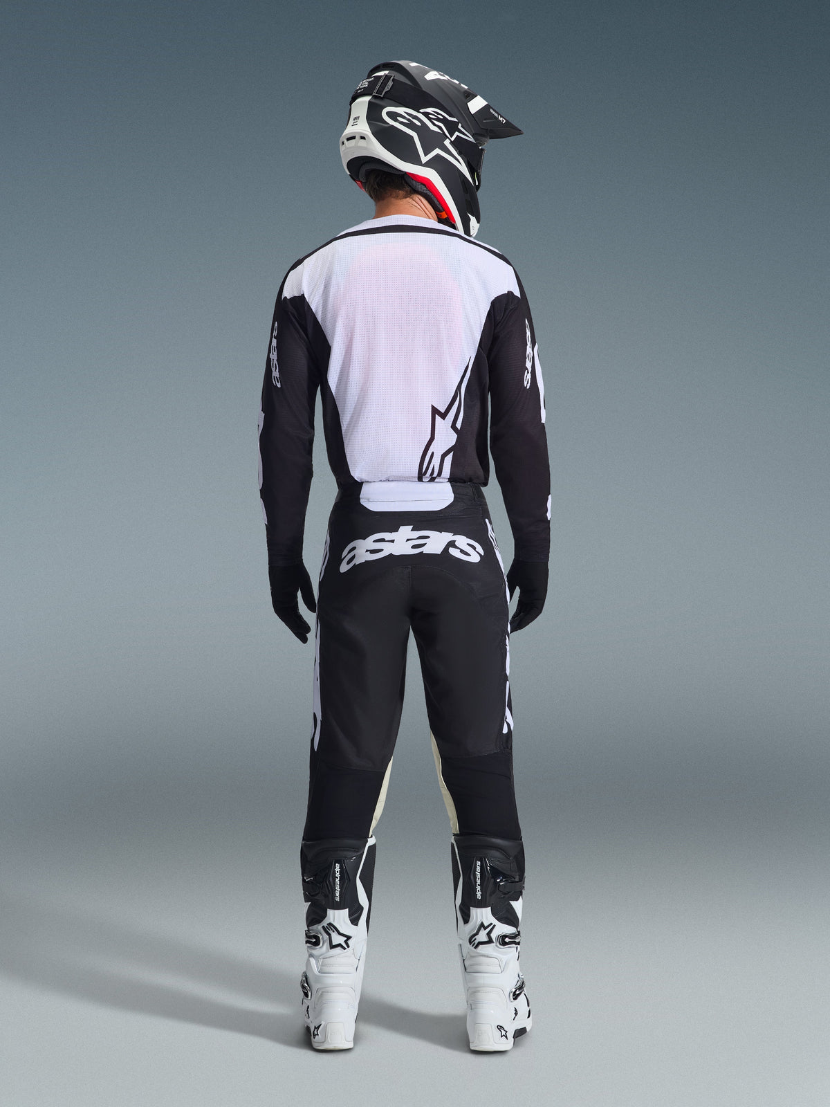 Pilota di motocross visto da dietro con pantaloni Alpinestars 2026 Racer Air Riway in nero e bianco, maglia coordinata a maniche lunghe nera e bianca, stivali da corsa bianchi e neri e casco nero con branding Alpinestars.