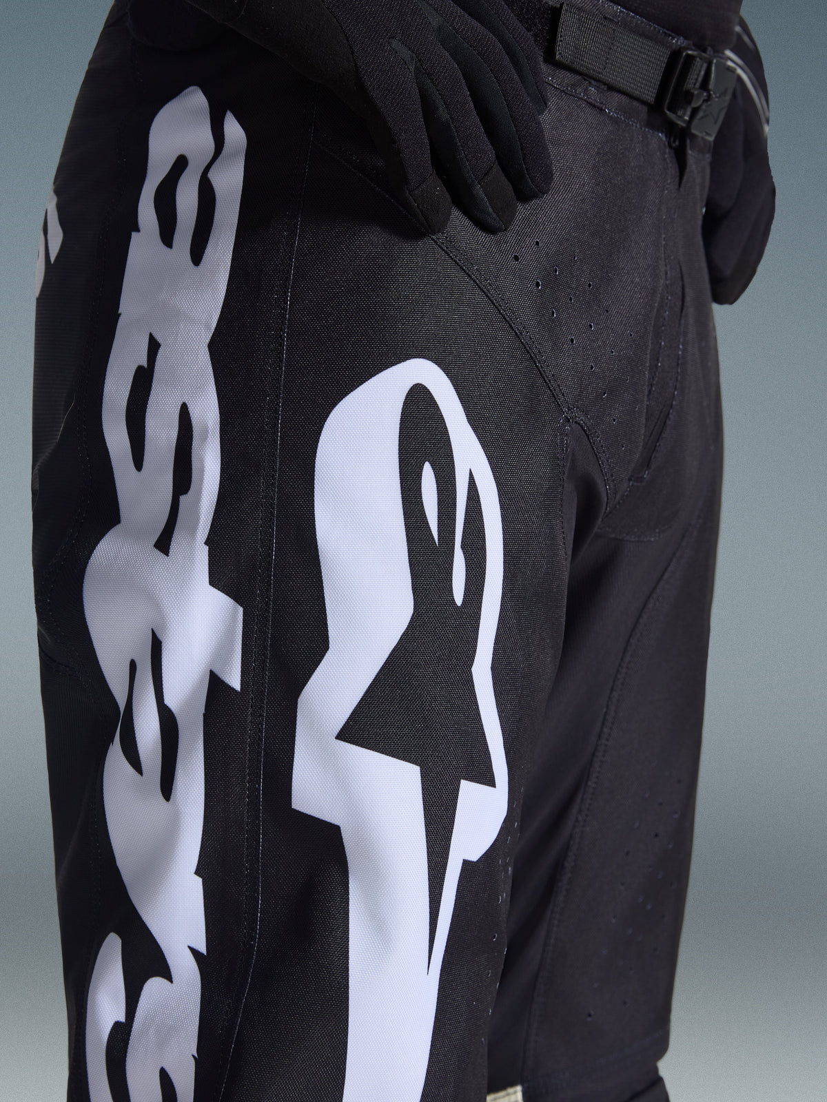Pantaloni Alpinestars Racer Air Riway 2026, pantaloni da motocross neri con grande logo Alpinestars bianco sulla coscia, primo piano del tessuto tecnico e della chiusura regolabile in vita, indossati con guanti neri per la guida fuoristrada