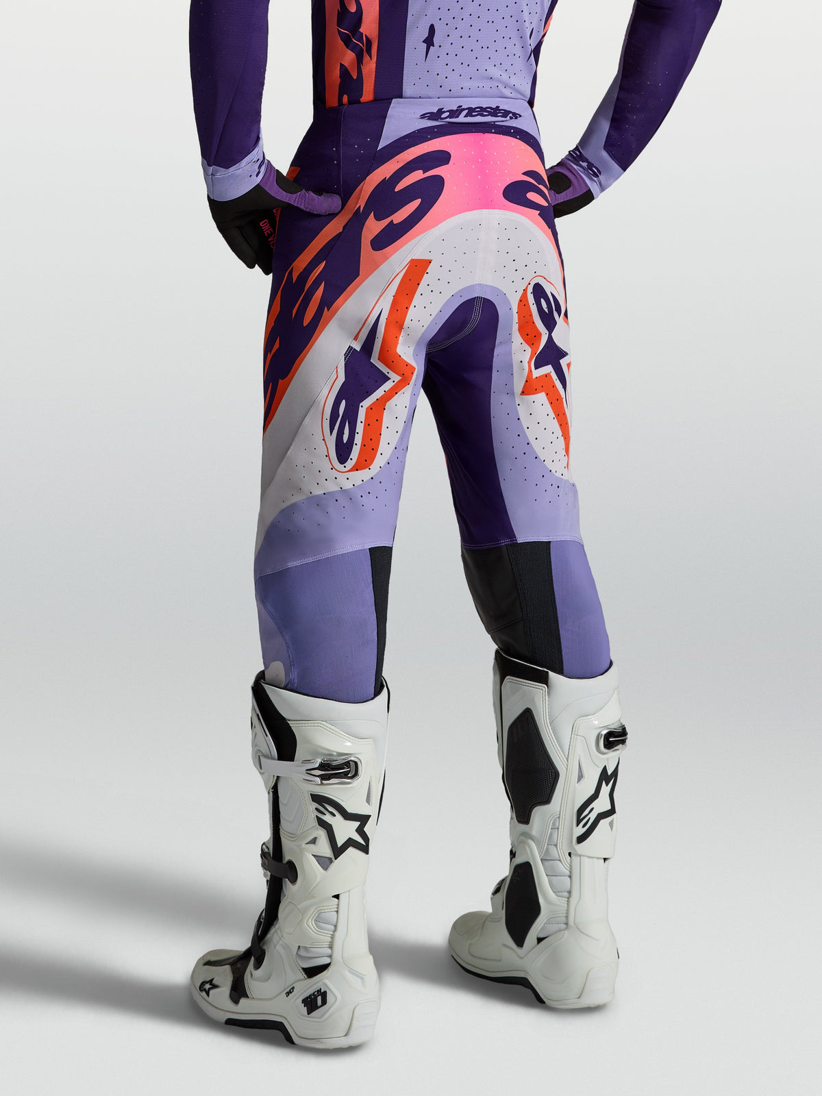 Alpinestars Pantaloni Supertech Dyna, pantaloni da motocross per uomo, viola e rosa fluo, in tessuto ripstop elastico e traspirante, vestibilità atletica, vista posteriore indossati con stivali bianchi.