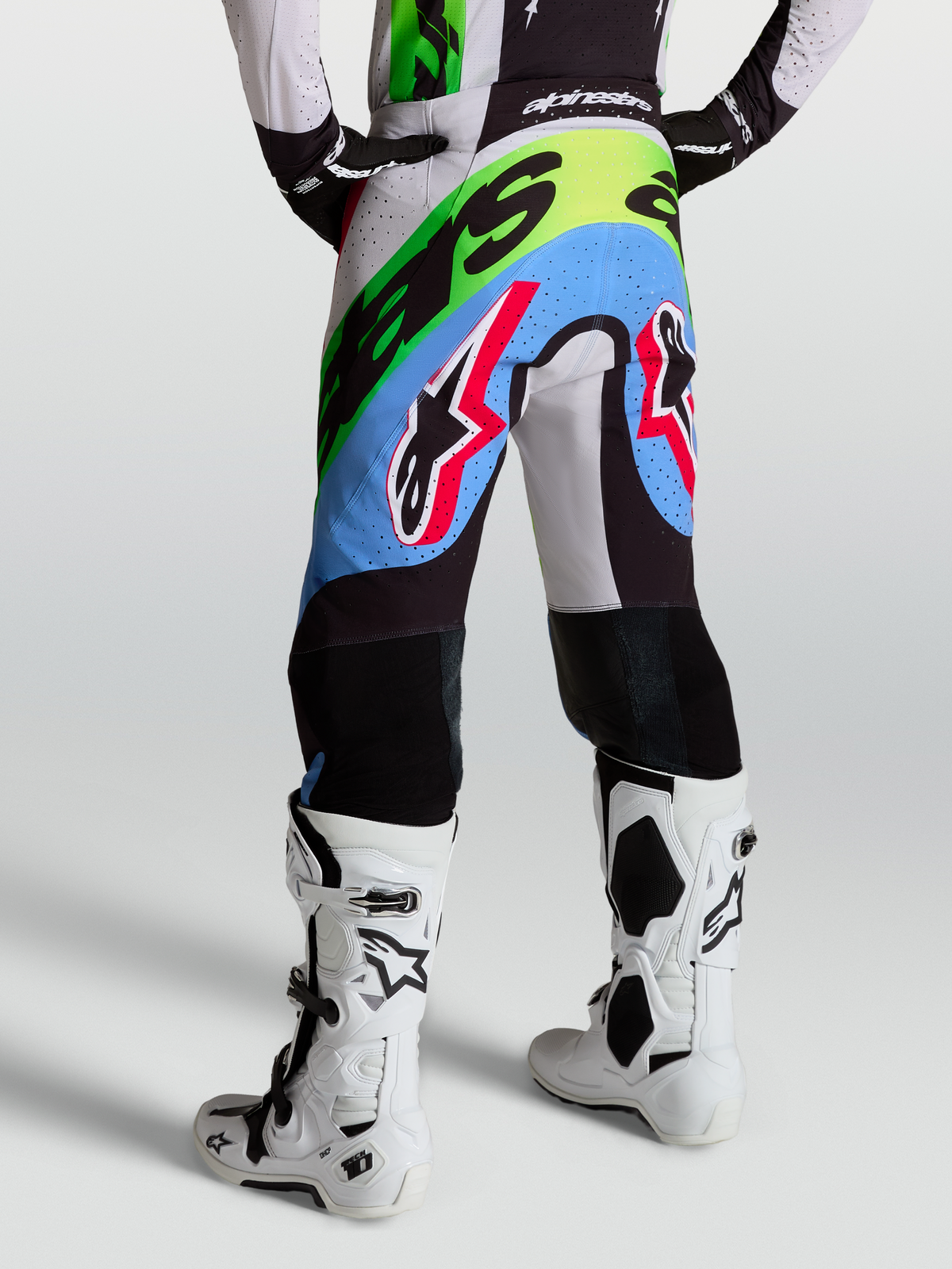 Alpinestars Pantaloni Supertech Dyna, pantaloni motocross da uomo, colore lilla chiaro, verde fluo e nero con rinforzi in pelle, indossati con stivali bianchi, abbigliamento racing off-road