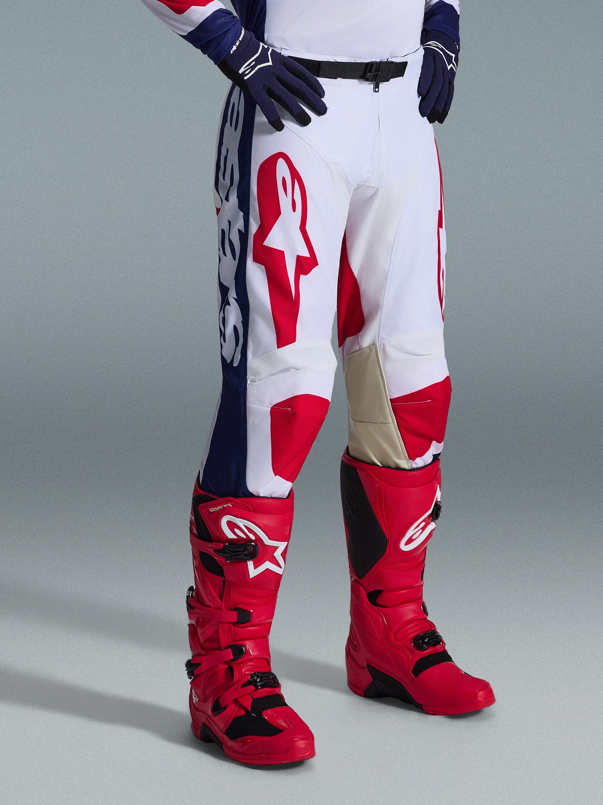 Alpinestars Pantaloni Racer Riway 2026, pantaloni da motocross, bianco blu rosso, con vistosi loghi rossi sulle cosce e pannelli laterali blu con marchio bianco, indossati con stivali rossi e guanti blu scuro, design ergonomico per corse fuoristrada