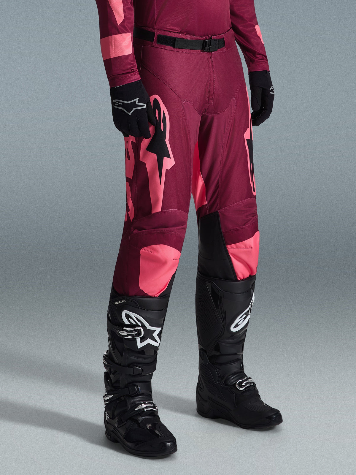Alpinestars 2026 Racer Riway Pants, pantaloni da motocross, Dark Red Coral, con grandi loghi a stella rosa e pannelli interni neri sulle ginocchia, indossati con stivali da motocross neri e guanti neri, vestibilità atletica da gara per il fuoristrada