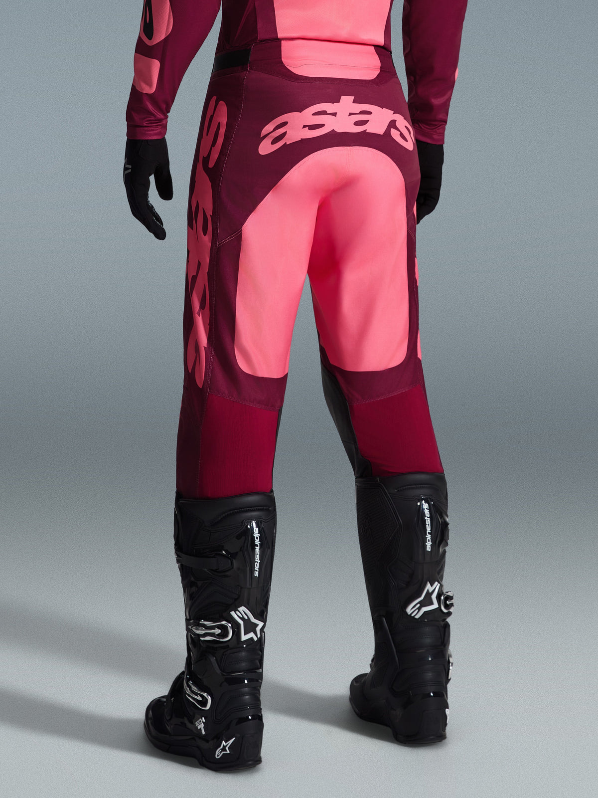 Alpinestars 2026 Racer Riway Pants, pantaloni da motocross, rosso scuro e corallo, vista posteriore con grande logo 'astars' color corallo sulla seduta, indossati con stivali e guanti da motocross neri, abbigliamento performante per corse fuoristrada