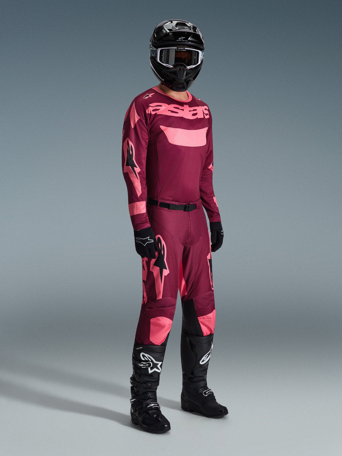 Pilota di motocross con abbigliamento completo da gara, Maglia Alpinestars 2026 Racer Riway in Dark Red Coral con dettagli rosa, pantaloni coordinati rosso scuro con loghi rosa, casco nero con occhiali dalla montatura bianca, guanti neri e stivali neri con loghi bianchi, completo coordinato per gare fuoristrada