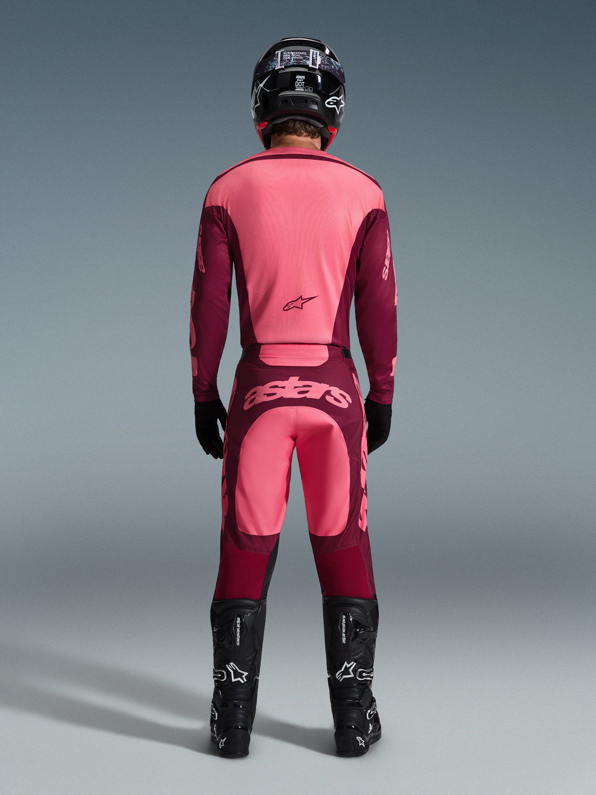 Pilota di motocross visto da dietro che indossa la maglia Alpinestars 2026 Racer Riway e i pantaloni coordinati in Dark Red Coral, con stivali neri, casco nero e guanti neri, equipaggiamento coordinato ad alte prestazioni per il fuoristrada