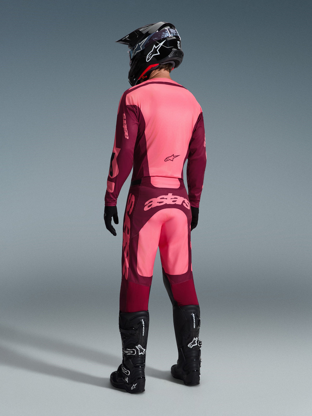 Pilota di motocross che indossa il completo Alpinestars 2026 Racer Riway visto da dietro, colorazione bordeaux e rosa chiaro, maglia e pantaloni con branding in evidenza, completo di casco, guanti e stivali neri, outfit da gara coordinato