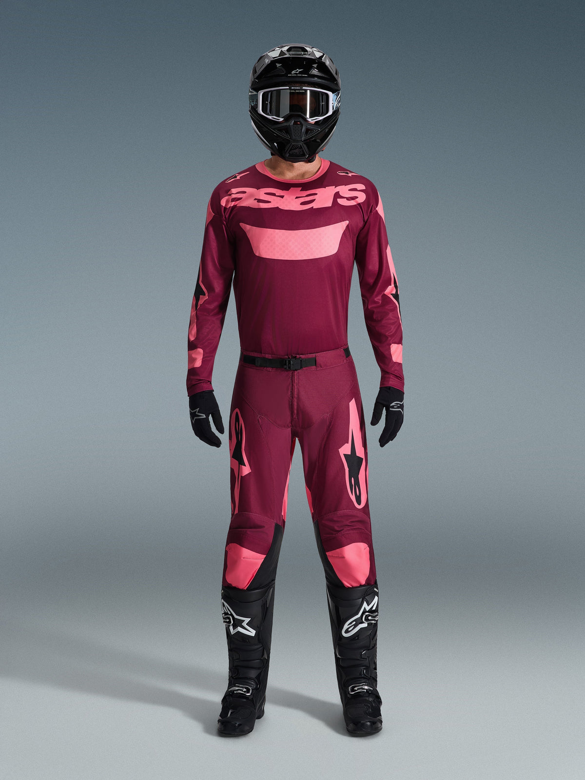 Pilota di motocross con abbigliamento completo da gara, maglia Dark Red Coral con accenti rosa, pantaloni Racer Riway coordinati, casco nero con dettagli bianchi, stivali neri con loghi bianchi, guanti neri, outfit coordinato per corse fuori strada