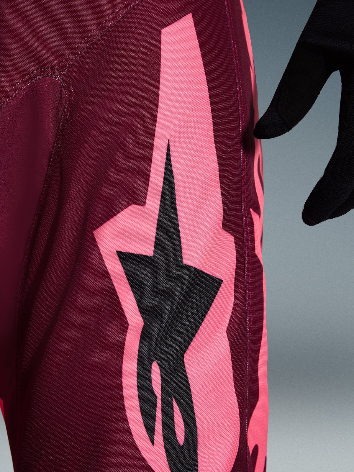 Pantaloni Alpinestars 2026 Racer Riway, pantaloni da motocross, Dark Red Coral, vista ravvicinata che mostra una grande grafica angolare rosa corallo e il logo a stella nero, mettendo in risalto la trama resistente in polyfabric e le cuciture tecniche