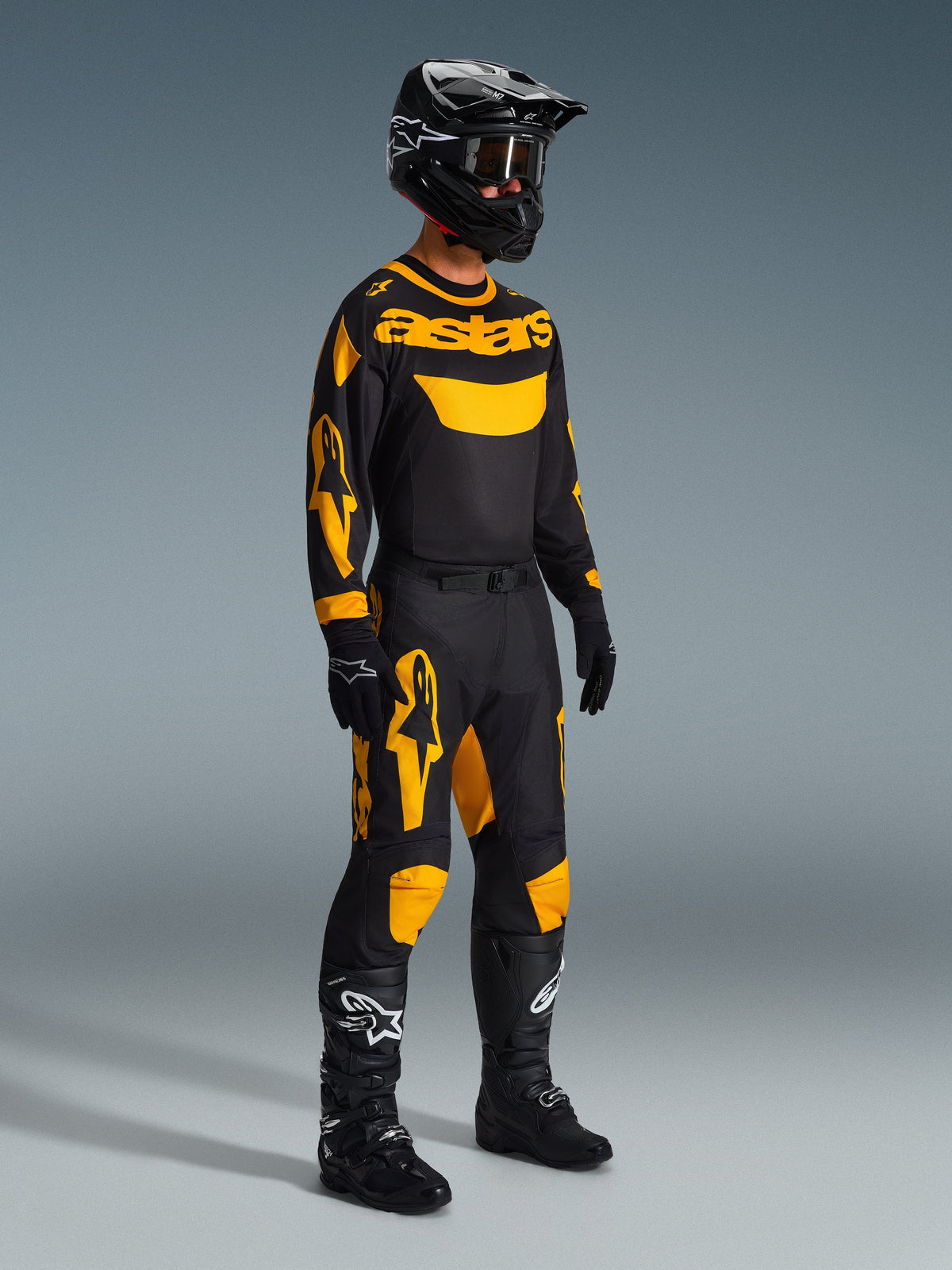 Pilota di motocross che indossa i pantaloni Alpinestars 2026 Racer Riway in giallo e nero, abbinati a una maglia 'astars' coordinata nera e gialla, casco nero con occhiali e stivali da gara neri, abbigliamento coordinato per prestazioni motocross off-road.