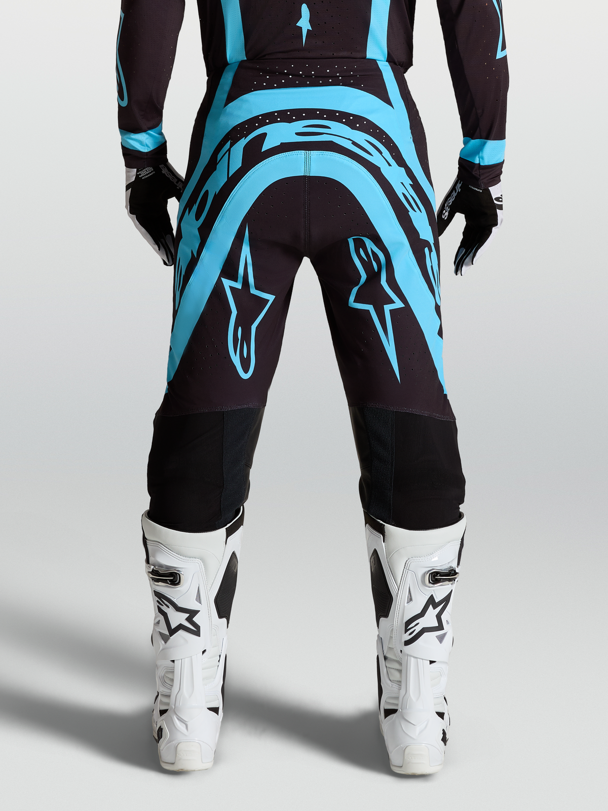 Alpinestars Pantaloni Supertech Vesta, pantaloni da motocross nero e turchese, vestibilità pro-level race-fit con stivali da moto bianchi, abbigliamento fuoristrada tecnico.
