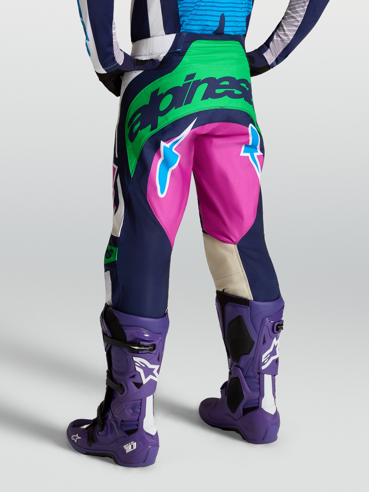 Alpinestars Pantaloni Techstar Maven, pantaloni motocross da uomo, blu e verde con dettagli fucsia, vista posteriore su modello con stivali Tech 10 viola.