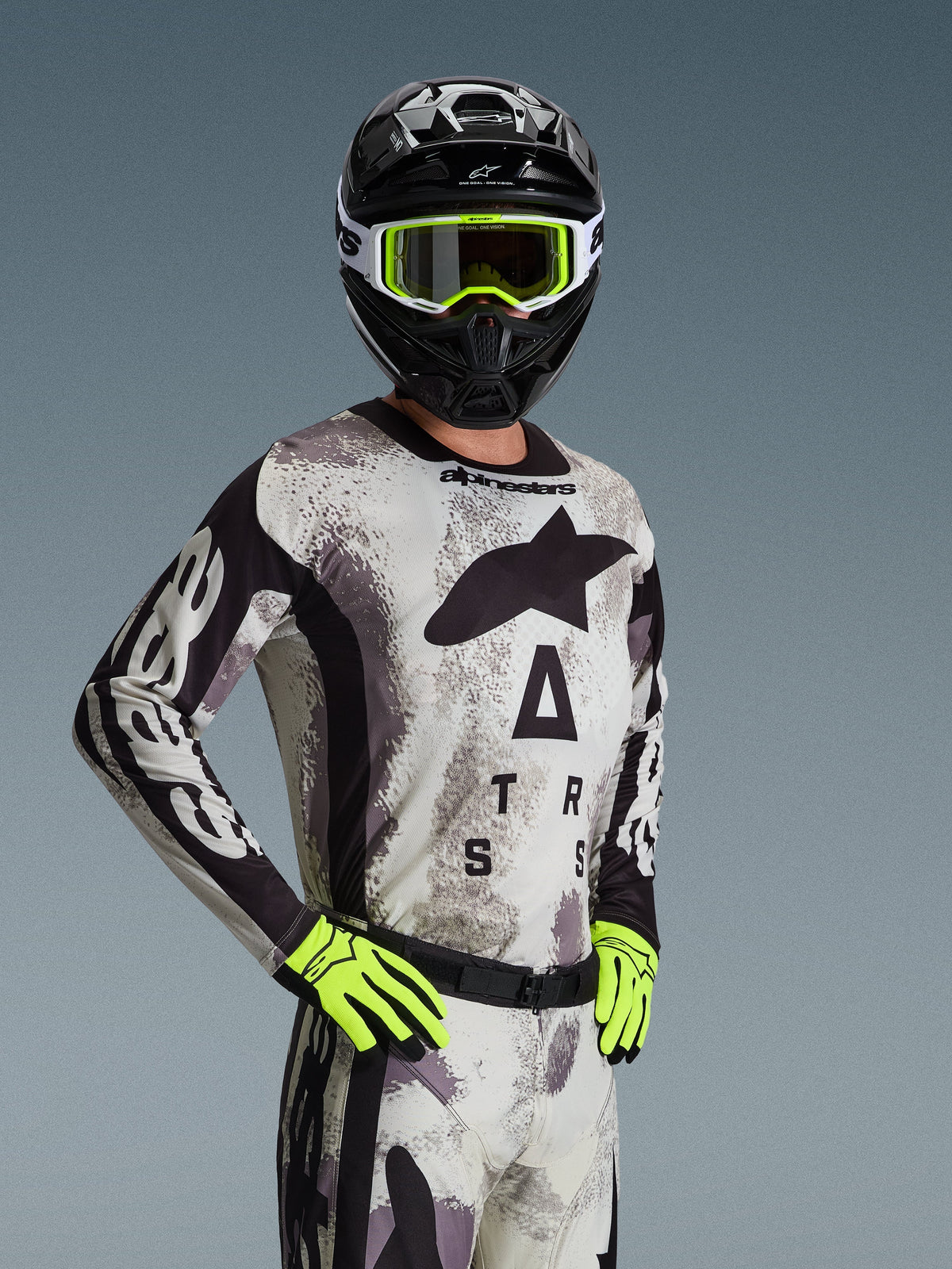 Pilota di motocross con maglia e completo Alpinestars Racer Lahnd 2026 in Sand Camo, motivo mimetico grigio chiaro con loghi neri, casco nero con occhiali bianchi e giallo neon, guanti giallo neon, abbigliamento professionale da fuoristrada
