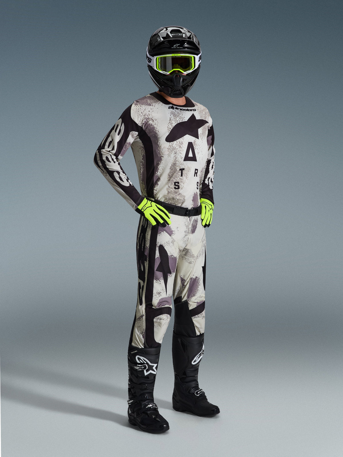 Alpinestars 2026 Racer Lahnd Gear Set, pilota di motocross con equipaggiamento completo da gara in Sand Camo e grigio, maglia e pantaloni coordinati con dettagli neri e loghi evidenti, maschera e guanti giallo neon, casco nero e stivali neri.