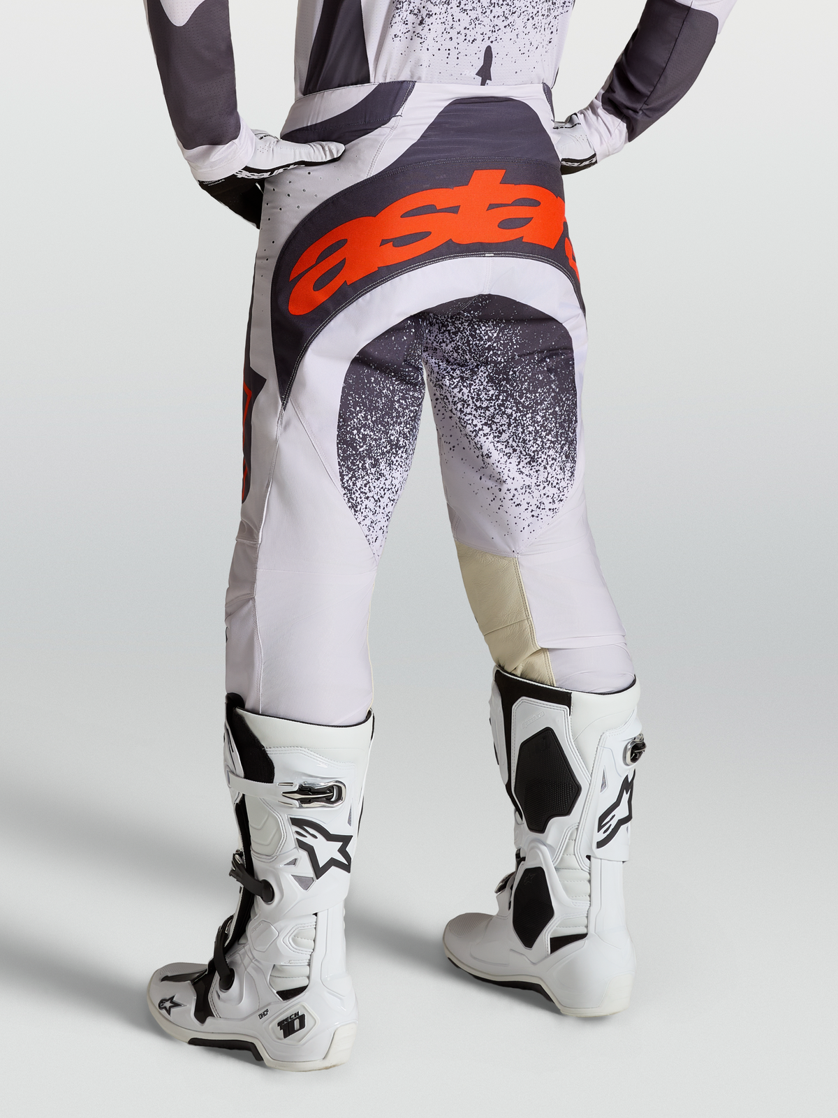 Alpinestars Pantaloni Techstar Swan, pantaloni da motocross grigi e lilla per uomo, con loghi rosso fluo e abbinati a stivali Tech 10 bianchi, vista posteriore su modello.