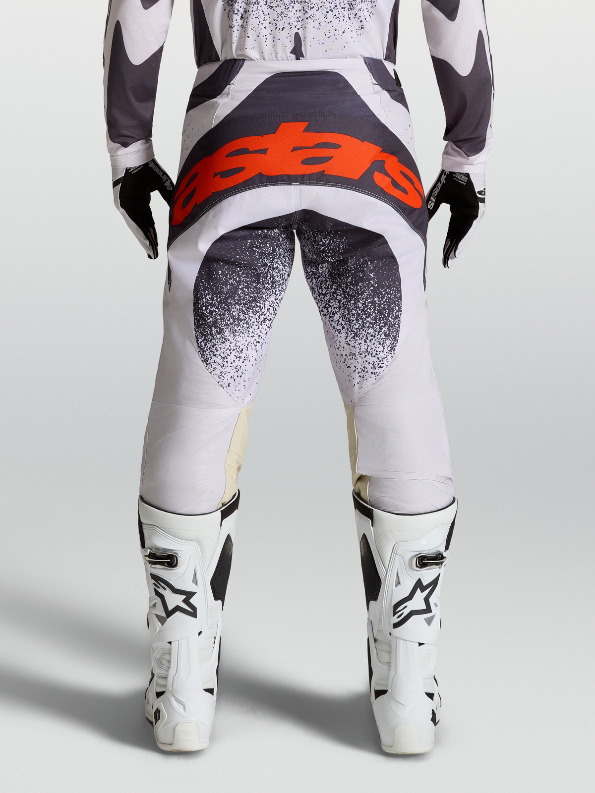 Alpinestars Pantaloni Techstar Swan, pantaloni da motocross racing uomo, grigio e lilla chiaro, logo Astars rosso fluo sul retro, indossati con stivali bianchi, abbigliamento fuoristrada performante
