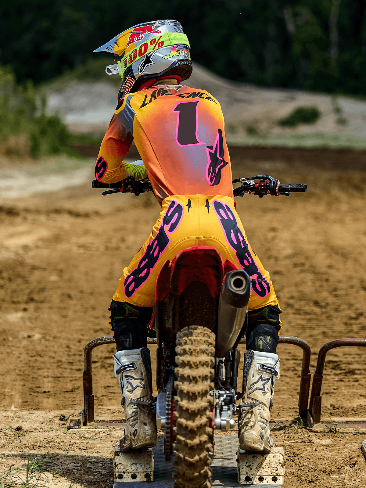 Pilota di motocross visto da dietro al cancelletto di partenza, indossa pantaloni Alpinestars Supertech Lipan 2025 in giallo e rosa, maglia coordinata con accenti rosa e numero 1, stivali Alpinestars bianchi e casco da gara, abbigliamento professionale coordinato per competizioni fuoristrada