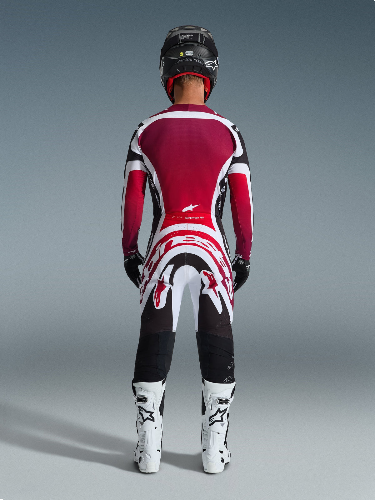 Pilota di motocross visto da dietro che indossa pantaloni Alpinestars 2026 Supertech AFD Vista, Nero Rosso Bianco, abbinati a una maglia coordinata, stivali da gara bianchi e casco nero, set completo di abbigliamento professionale per gare fuoristrada