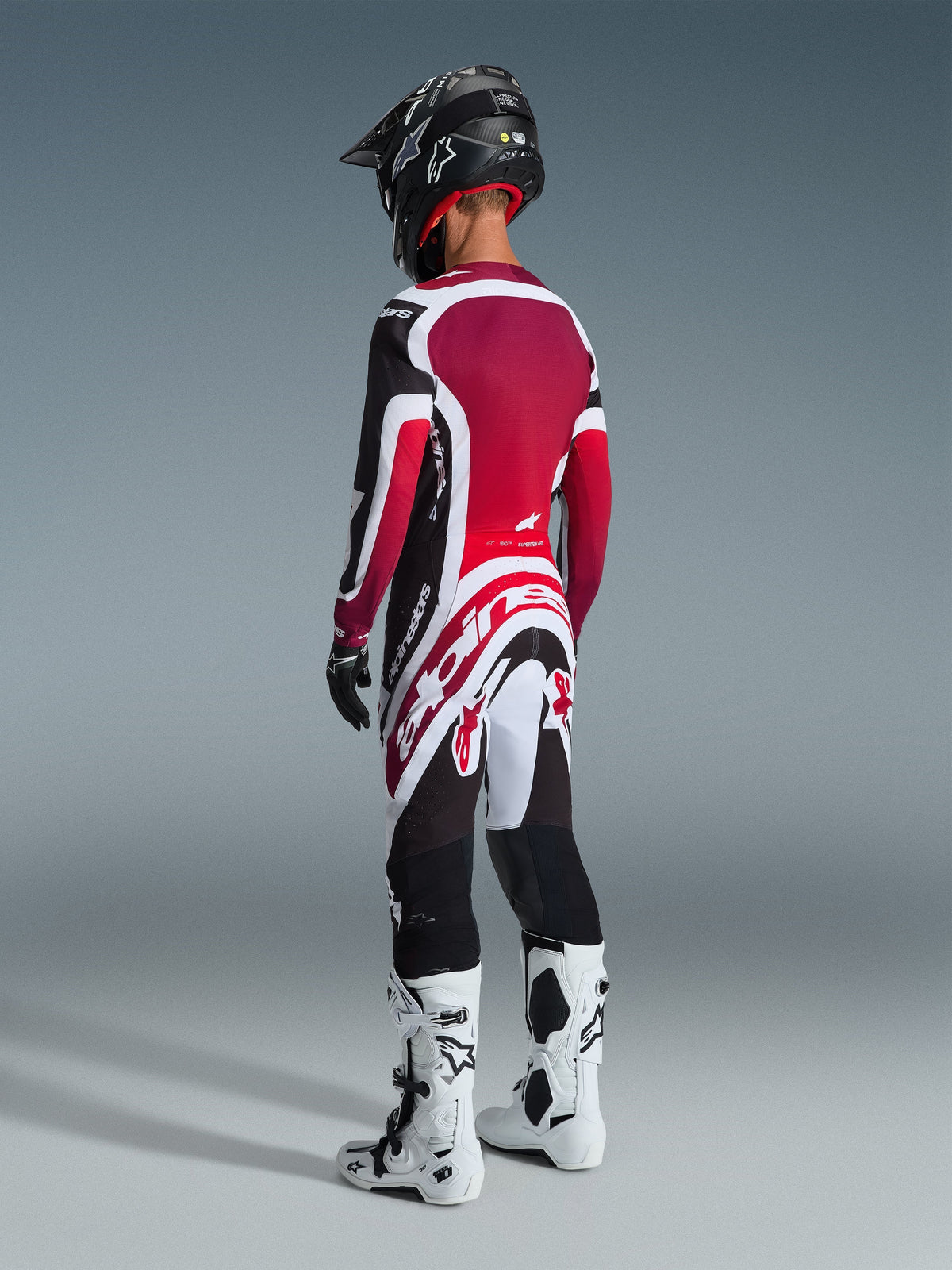 Pilota di motocross visto da dietro con pantaloni Alpinestars 2026 Supertech AFD Vista, nero, rosso e bianco, maglia coordinata, casco nero e stivali bianchi e neri, vista posteriore a figura intera di abbigliamento da gara professionale