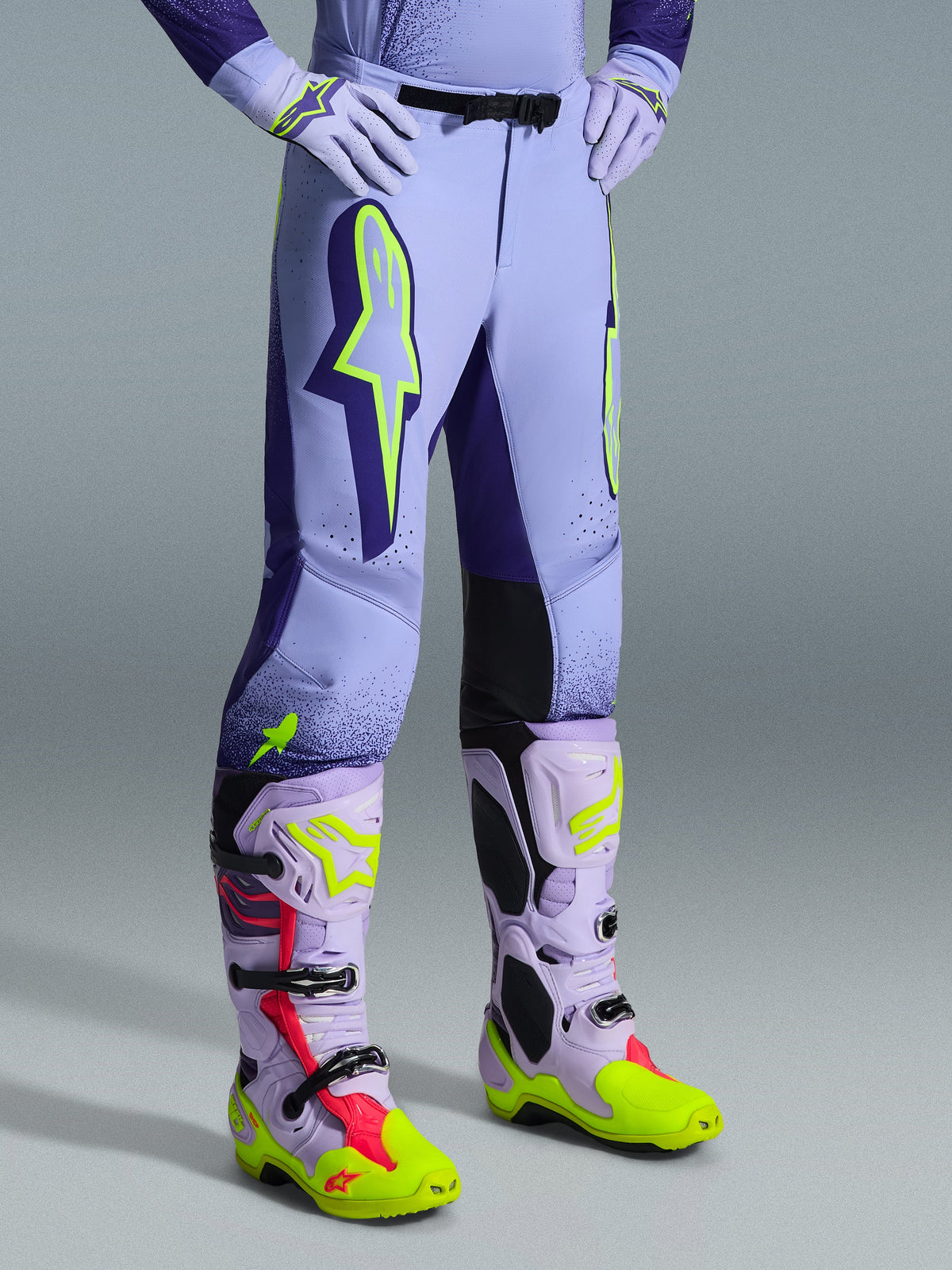 Alpinestars 2026 Supertech Scenz Pants, pantaloni da motocross, viola e giallo fluo, indossati con stivali e guanti da motocross coordinati, caratterizzati da grandi loghi giallo neon, vestibilità atletica e perforazioni tagliate al laser per la ventilazione