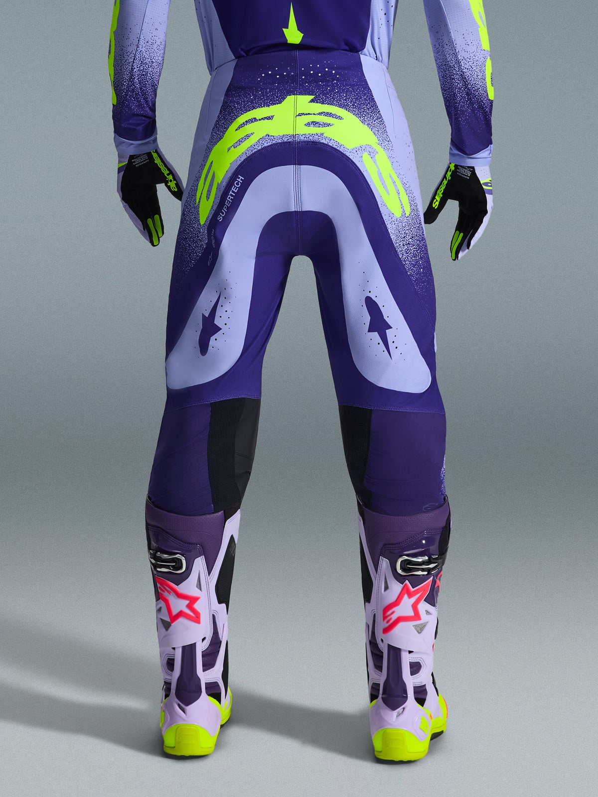 Alpinestars 2026 Supertech Scenz Pants, pantaloni da motocross, viola e giallo fluo, vista posteriore con grande logo giallo sulla seduta, indossati con maglia, guanti e stivali protettivi coordinati, vestibilità atletica per gare di motocross professionistiche
