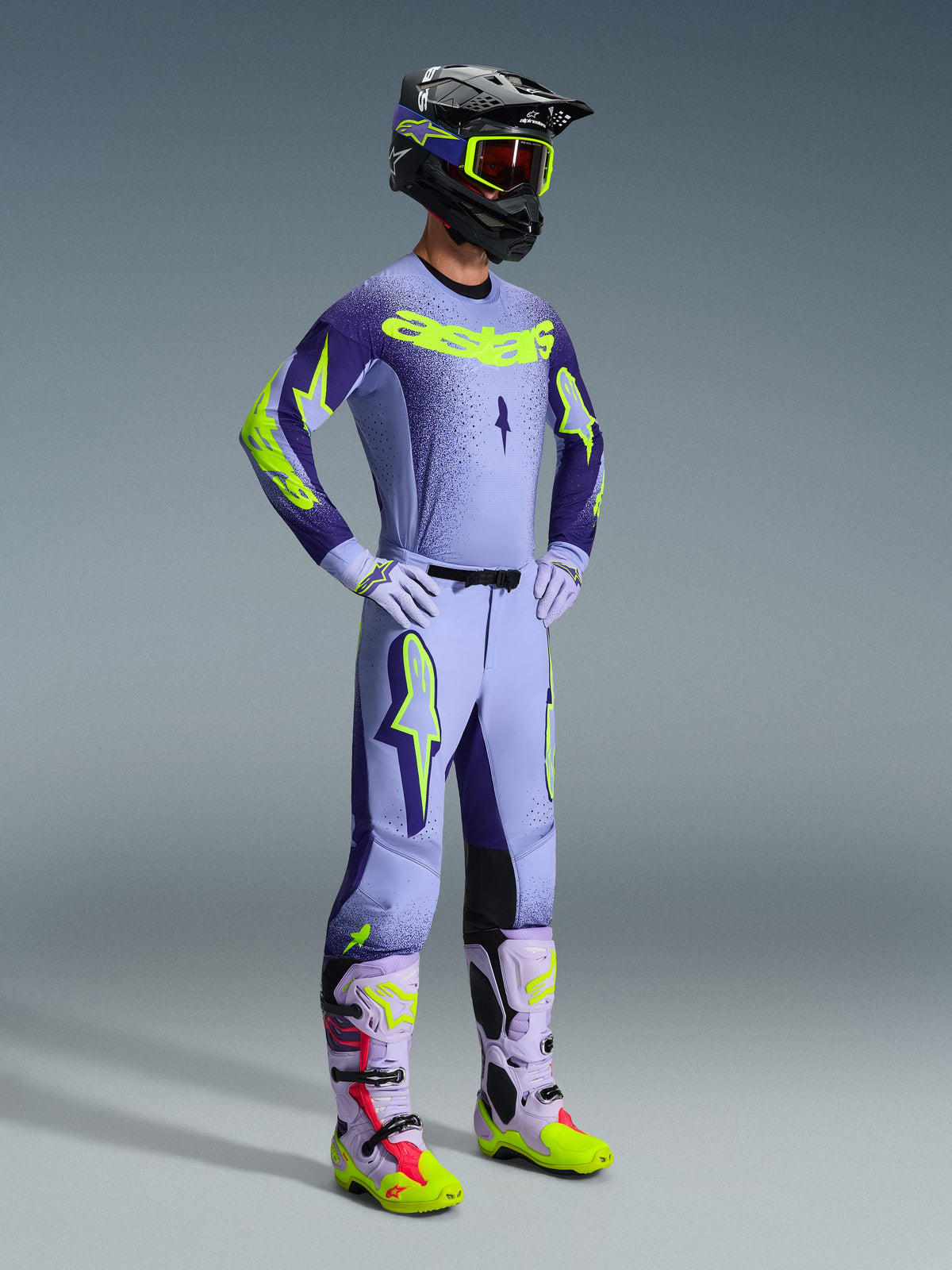 Alpinestars 2026 Supertech Scenz Pants, pantaloni da motocross, viola e giallo fluo, indossati da un pilota in un completo da gara con maglia sfumata coordinata, guanti lilla e stivali coordinati gialli e viola, con casco nero e occhiali.