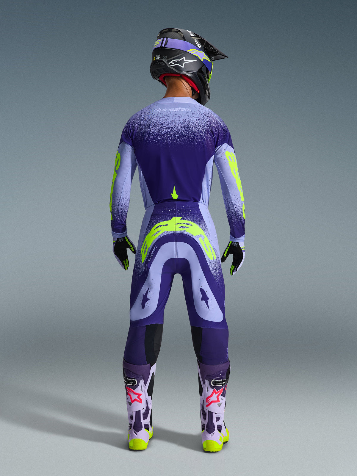 Pilota di motocross visto da dietro che indossa i pantaloni Alpinestars 2026 Supertech Scenz in Purple Yellow Fluo, maglia coordinata con grafiche sfumate viola e giallo neon, casco nero e stivali viola, abbigliamento da gara professionale coordinato per prestazioni fuoristrada