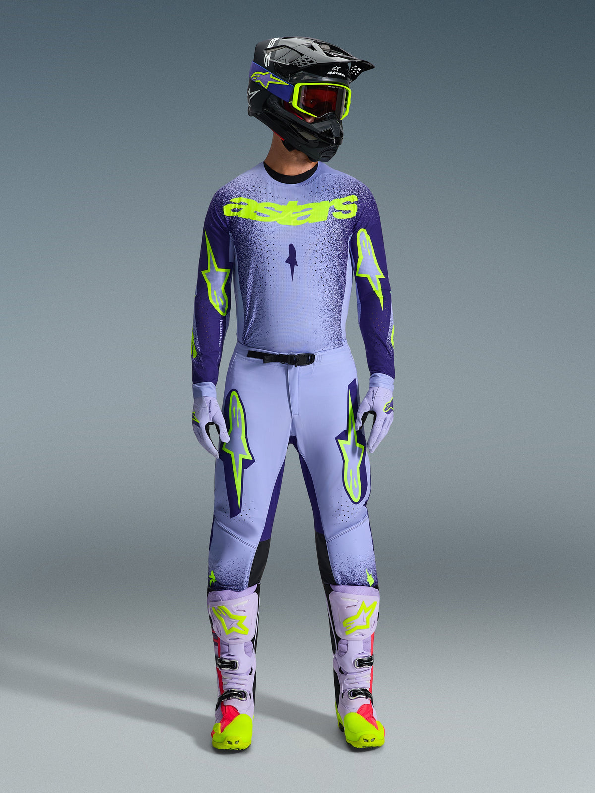 Alpinestars Pantaloni Supertech Scenz 2026, pantaloni da motocross viola e giallo fluo, indossati come completo da gara con maglia, guanti e stivali lavanda coordinati, caratterizzati da vivaci loghi giallo fluo e design con vestibilità atletica per gare off-road professionali.