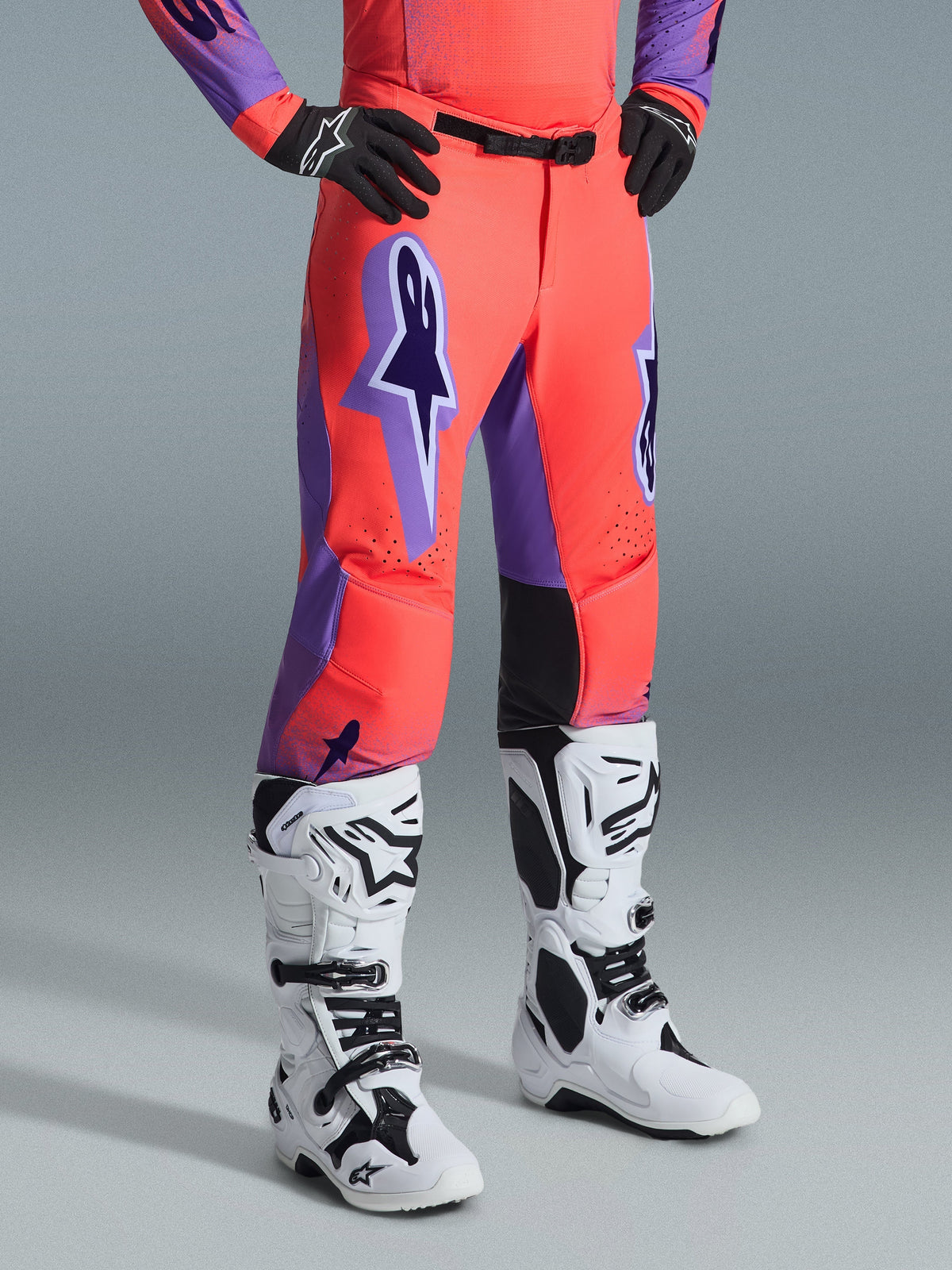 Alpinestars 2026 Supertech Scenz Pants, pantaloni da motocross, Hot Coral e viola, indossati con stivali da motocross bianchi e neri e guanti neri, con un grande logo Alpinestars viola, pannelli interni al ginocchio in pelle nera e design con vestibilità atletica da gara