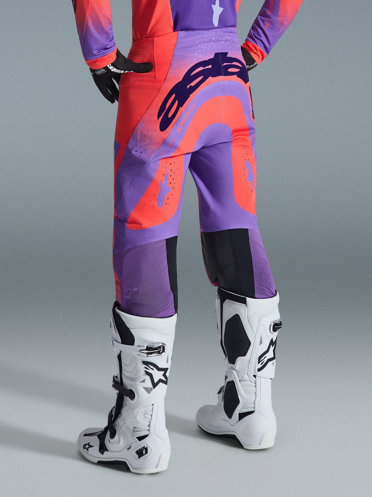 Alpinestars Pantaloni Supertech Scenz 2026, pantaloni da motocross, color corallo acceso e viola, vista posteriore con logo evidente sulla seduta, pannelli vivaci corallo e viola con zone traforate per la ventilazione, indossati con stivali da gara bianchi e neri