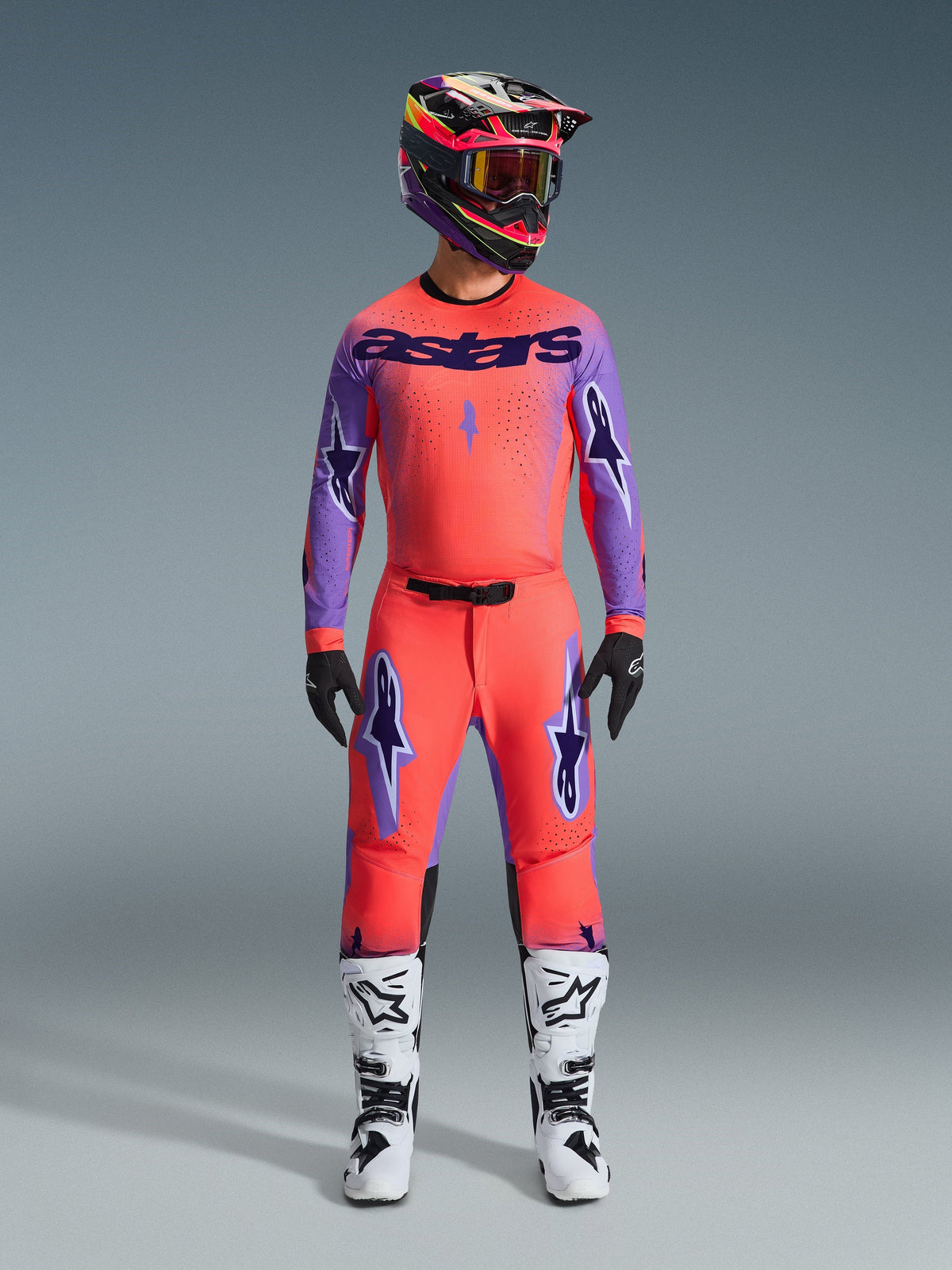 Pantaloni Alpinestars 2026 Supertech Scenz, pantaloni da motocross, corallo acceso e viola, indossati come set completo con maglia e casco coordinati, stivali bianchi e neri, con loghi viola e vestibilità atletica racing.