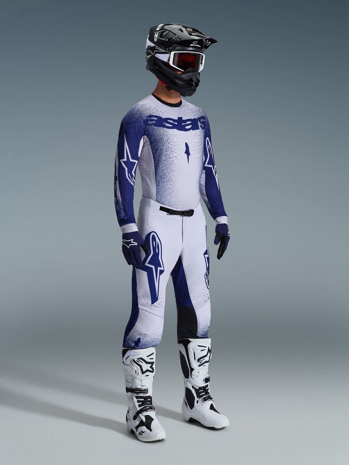 Pilota di motocross con pantaloni Supertech Scenz 2026 in grigio chiaro e blu scuro, maglia blu e bianca coordinata con logo 'astars' ed effetto spray, casco nero con occhiali bianchi, stivali da motocross bianchi e neri, completo professionale da gara fuoristrada.