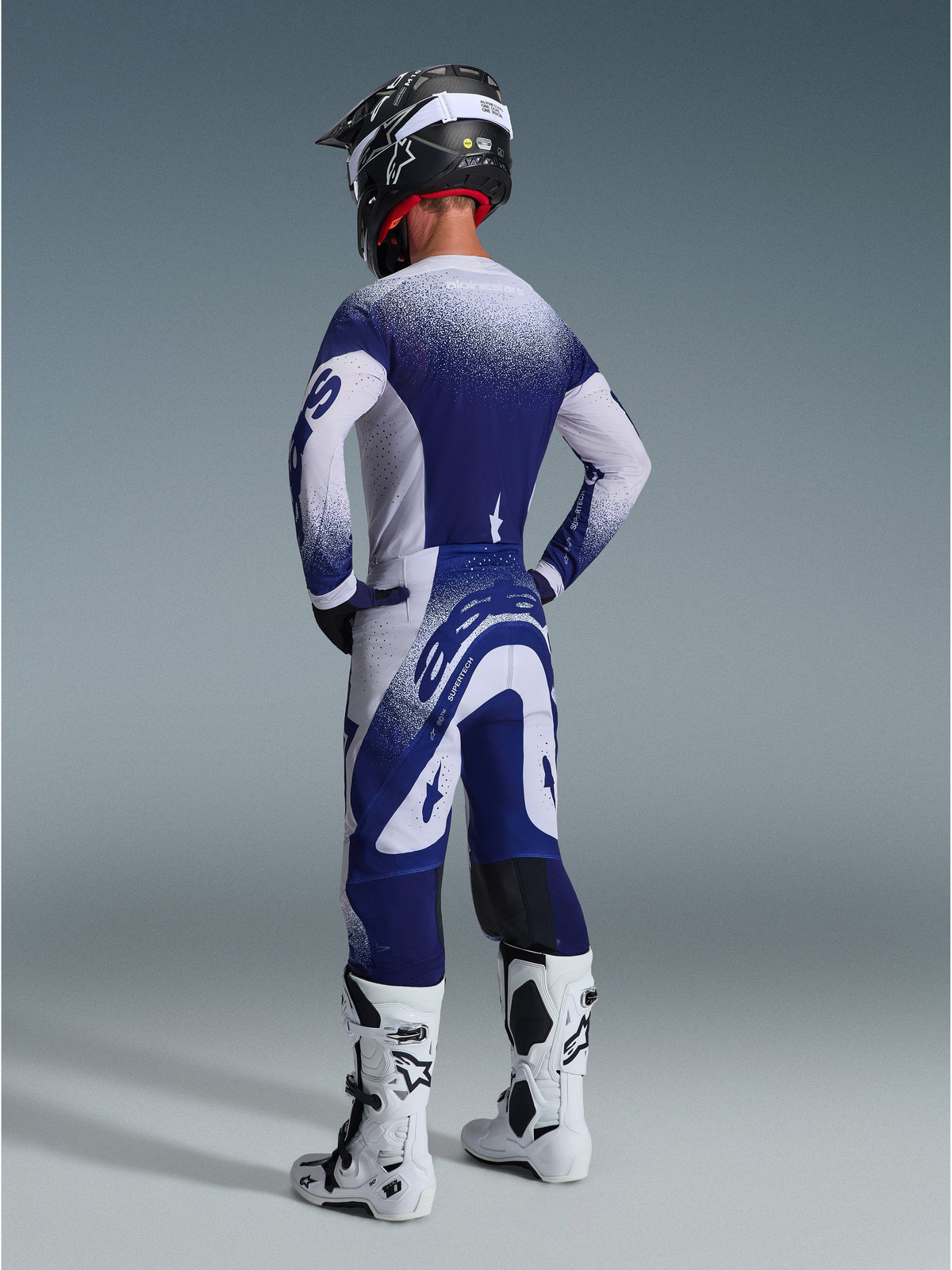 Alpinestars 2026 Supertech Scenz Pants, pantaloni da motocross, grigio chiaro e blu scuro, vista posteriore indossati con maglia coordinata e stivali bianchi, con grafiche a puntini sfumati e vestibilità atletica per gare fuoristrada