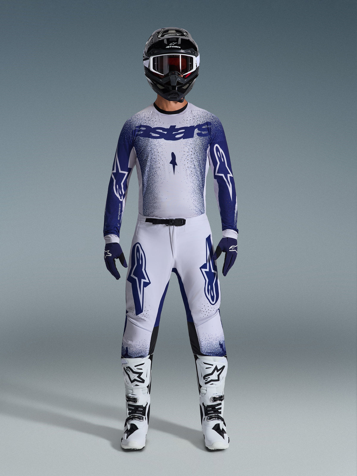 Pilota di motocross che indossa il completo Alpinestars Supertech Scenz 2026, grigio chiaro e blu scuro, composto da maglia e pantaloni con guanti blu coordinati, stivali bianchi e neri e casco nero con occhiali bianchi, outfit coordinato per gare professionali off-road