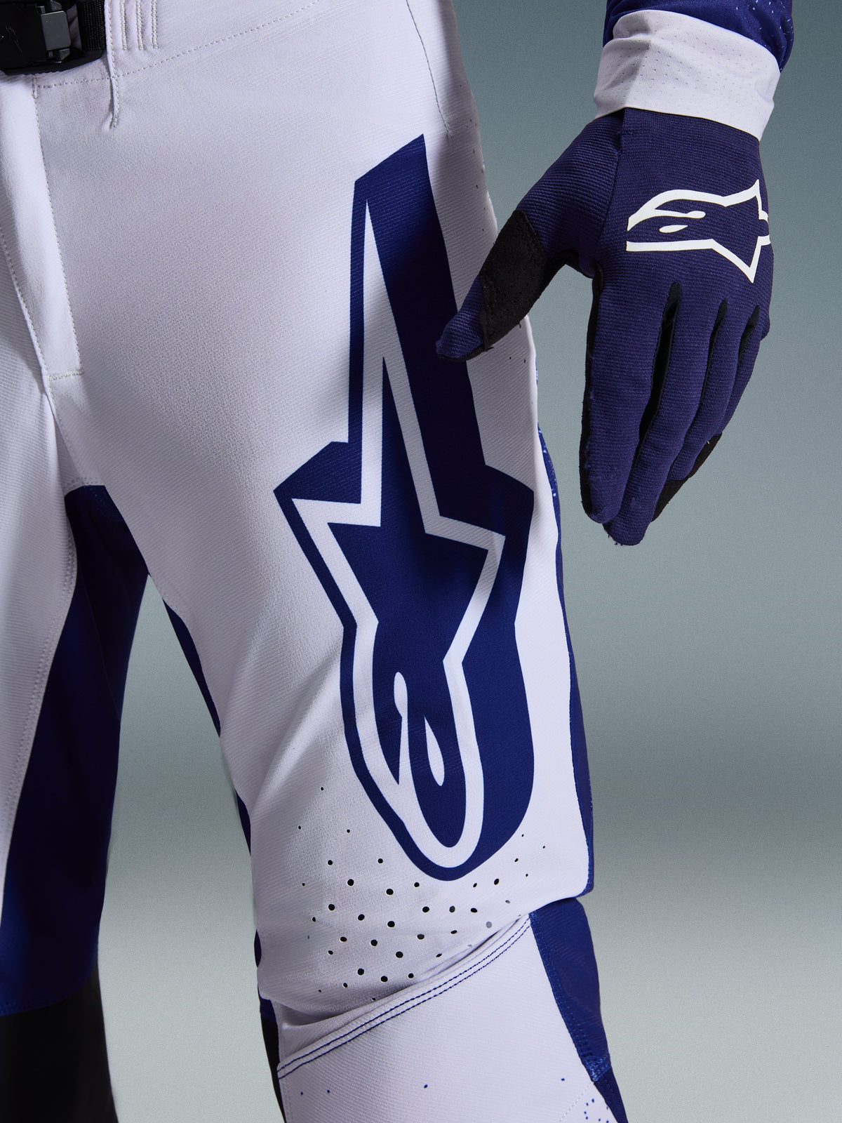 Alpinestars Pantaloni Supertech Scenz 2026, pantaloni da motocross, grigio chiaro e blu scuro, vista ravvicinata della coscia con grande logo Alpinestars blu scuro, zone di ventilazione traforate e tessuto rip-stop elasticizzato, indossati con un guanto da motocross blu scuro coordinato