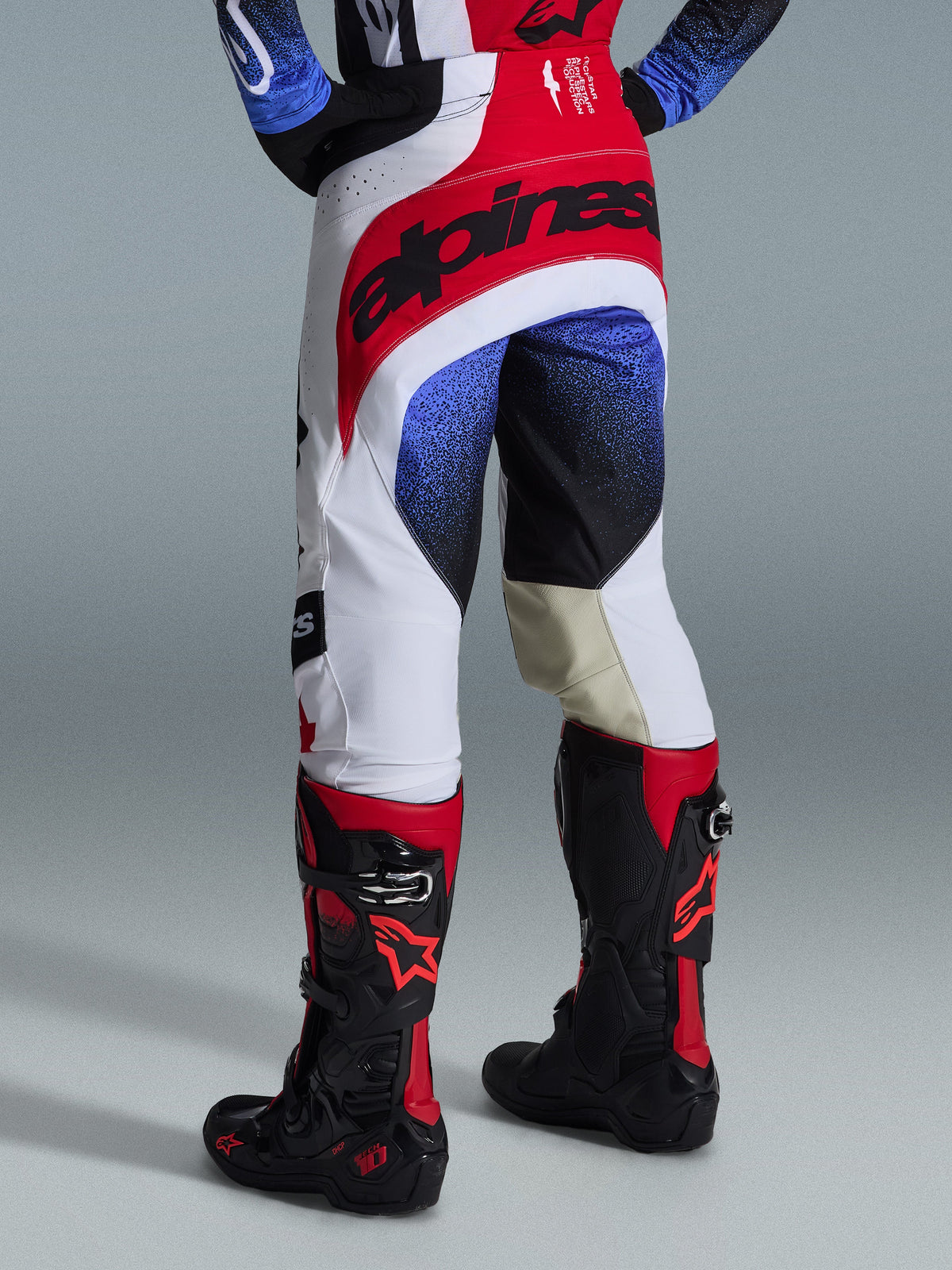 Alpinestars 2026 Techstar Knif Pants, pantaloni da motocross, nero, bianco e rosso con accenti blu, vista posteriore con logo evidente sulla vita e pannelli interni alle ginocchia in pelle color cuoio, indossati con stivali neri e rossi, abbigliamento tecnico ad alte prestazioni per competizioni off-road