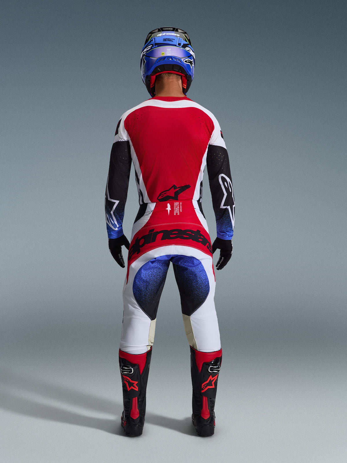 Alpinestars 2026 Techstar Knif Pants, pantaloni da motocross, nero bianco rosso e blu, vista posteriore di un pilota in equipaggiamento completo, maglia coordinata, casco blu e stivali neri e rossi, con vistoso marchio Alpinestars sulla seduta.