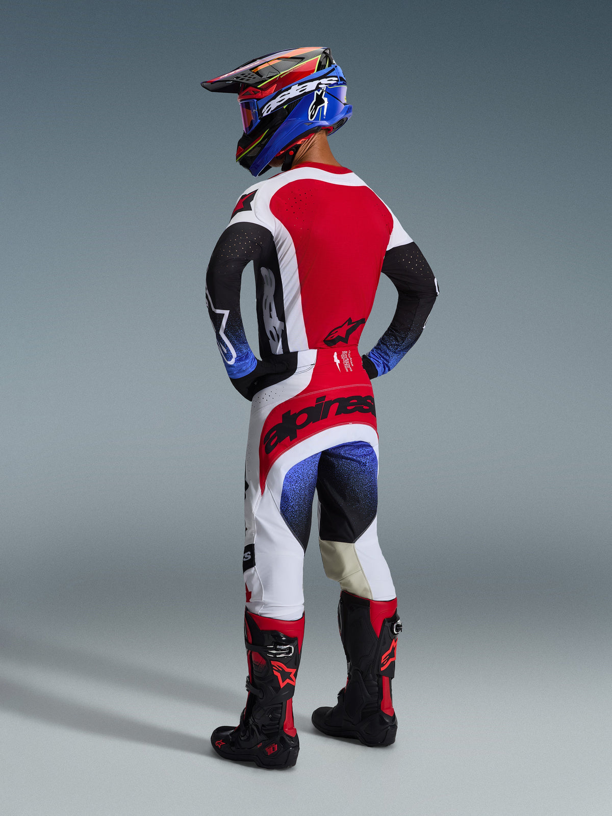 Alpinestars Pantaloni Techstar Knif 2026, pantaloni da motocross, nero, bianco e rosso con accenti sfumati blu, vista posteriore del pilota con equipaggiamento completo, logo rosso sulla seduta, pannelli bianchi e neri, indossati con maglia coordinata e stivali Tech 10
