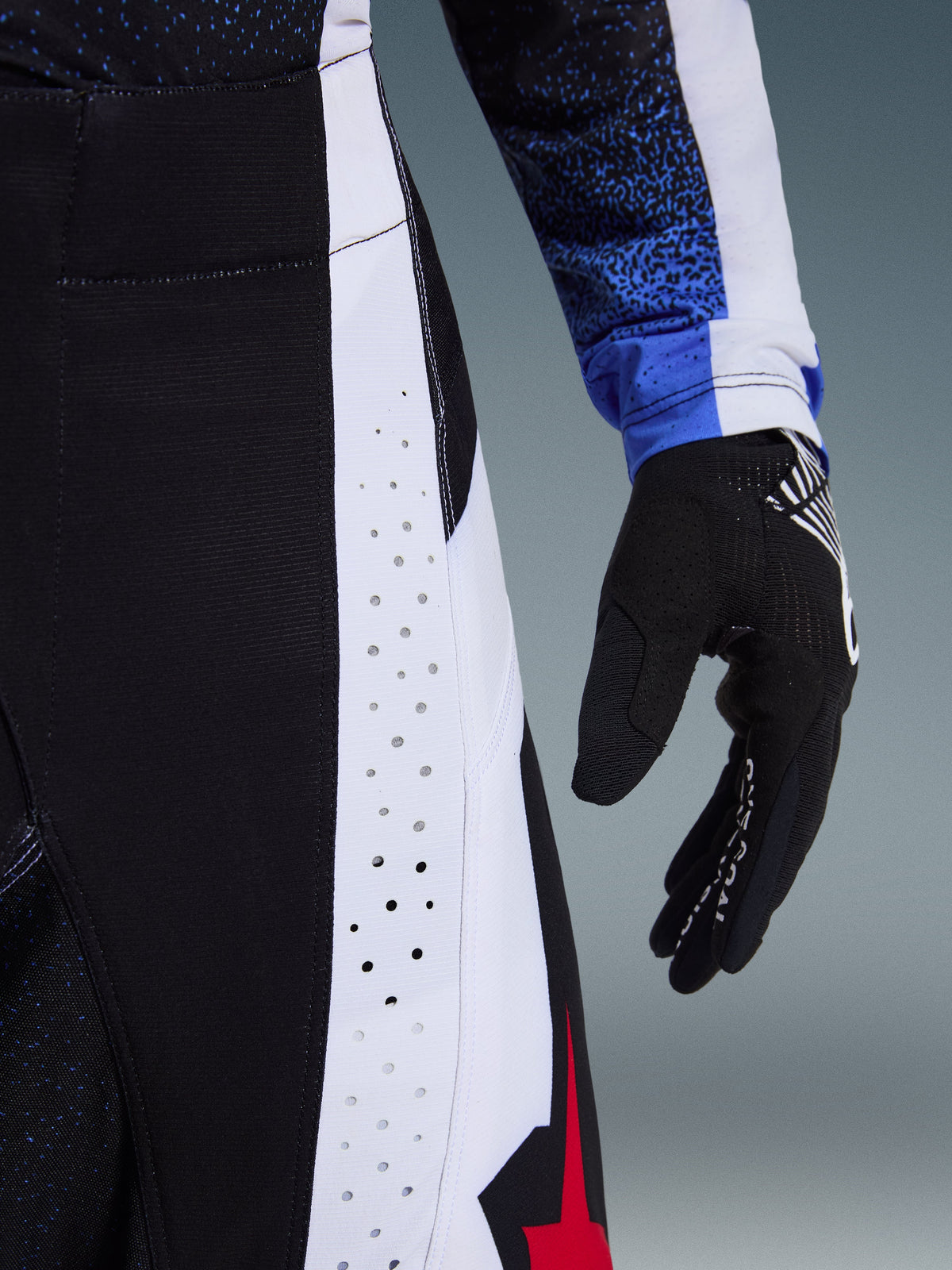 Alpinestars 2026 Techstar Knif Pants, pantaloni da motocross, nero, bianco e rosso, primo piano che evidenzia le perforazioni al laser sui pannelli laterali e il logo a stella rosso, abbinati a una maglia coordinata e a un guanto da motocross nero.