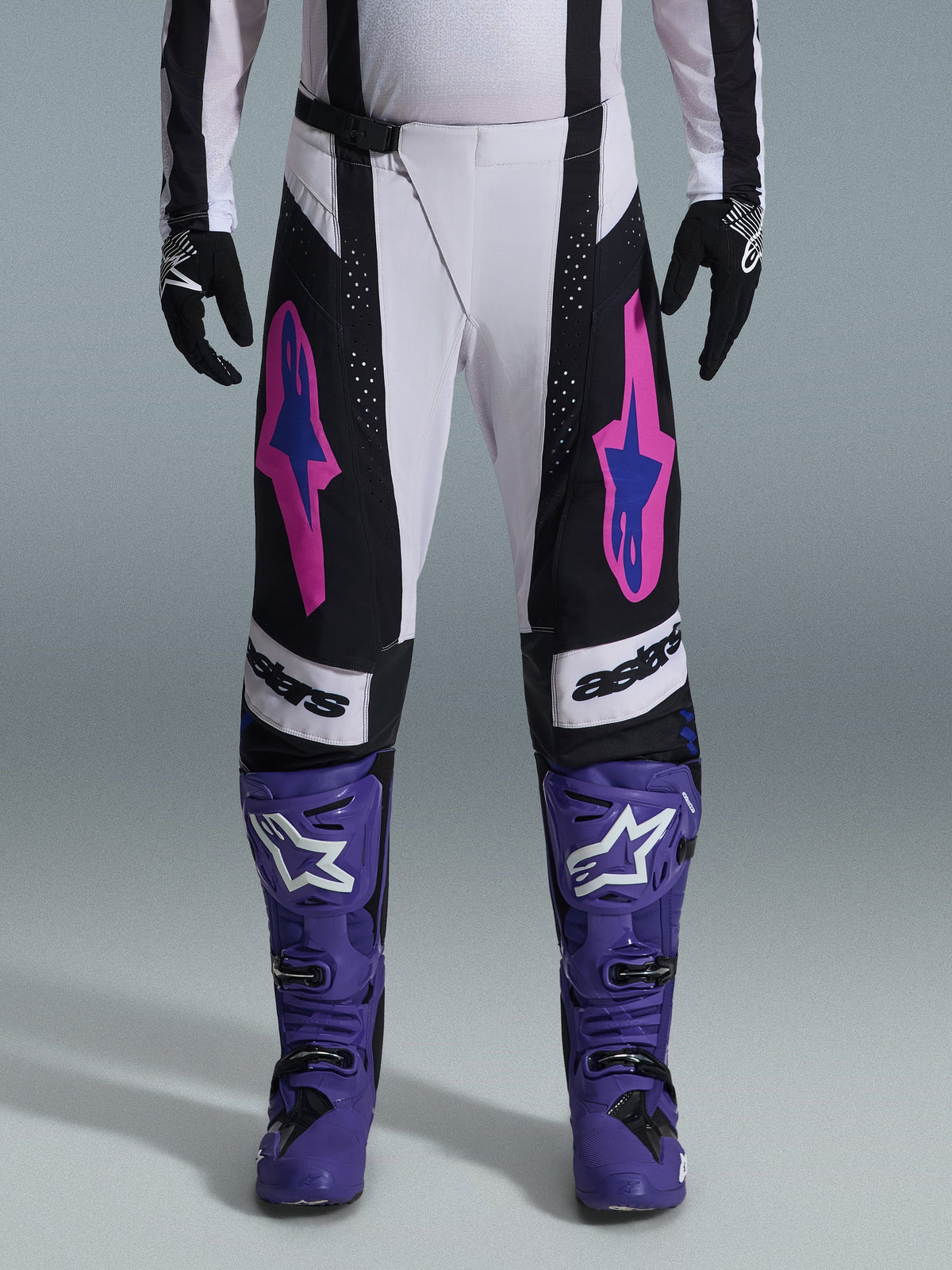 Pilota di motocross che indossa il completo Alpinestars Techstar Knif 2026, pantaloni bianchi e neri con loghi rosa e blu, stivali viola con marchio bianco, guanti neri, vestibilità race atletica e ventilazione tagliata al laser per gare fuoristrada