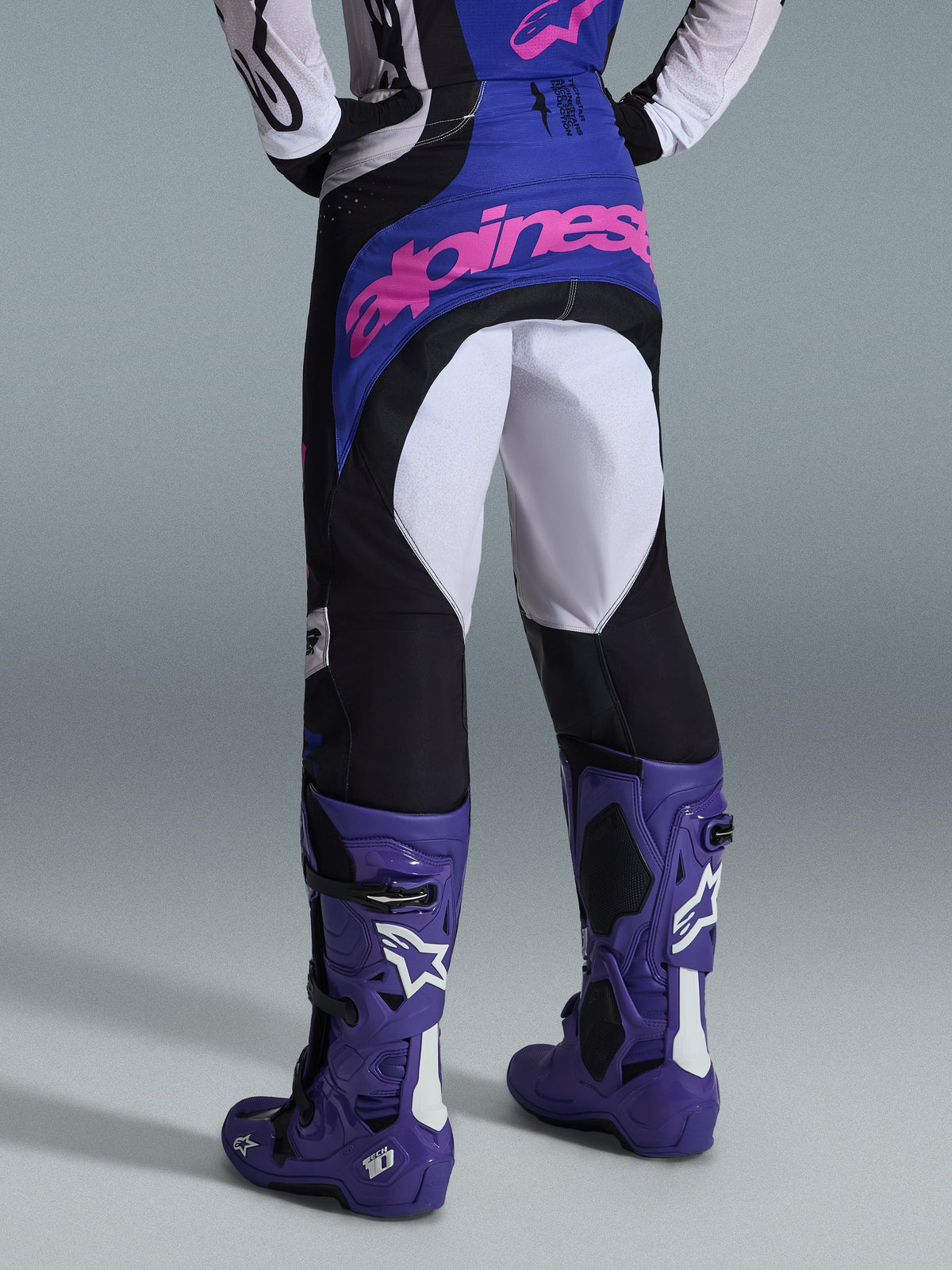Alpinestars Techstar Knif 2026, pantaloni da motocross, nero, grigio chiaro e viola, vista posteriore con logo rosa sulla seduta, indossati con maglia coordinata e stivali da motocross viola, completo professionale da gara off-road