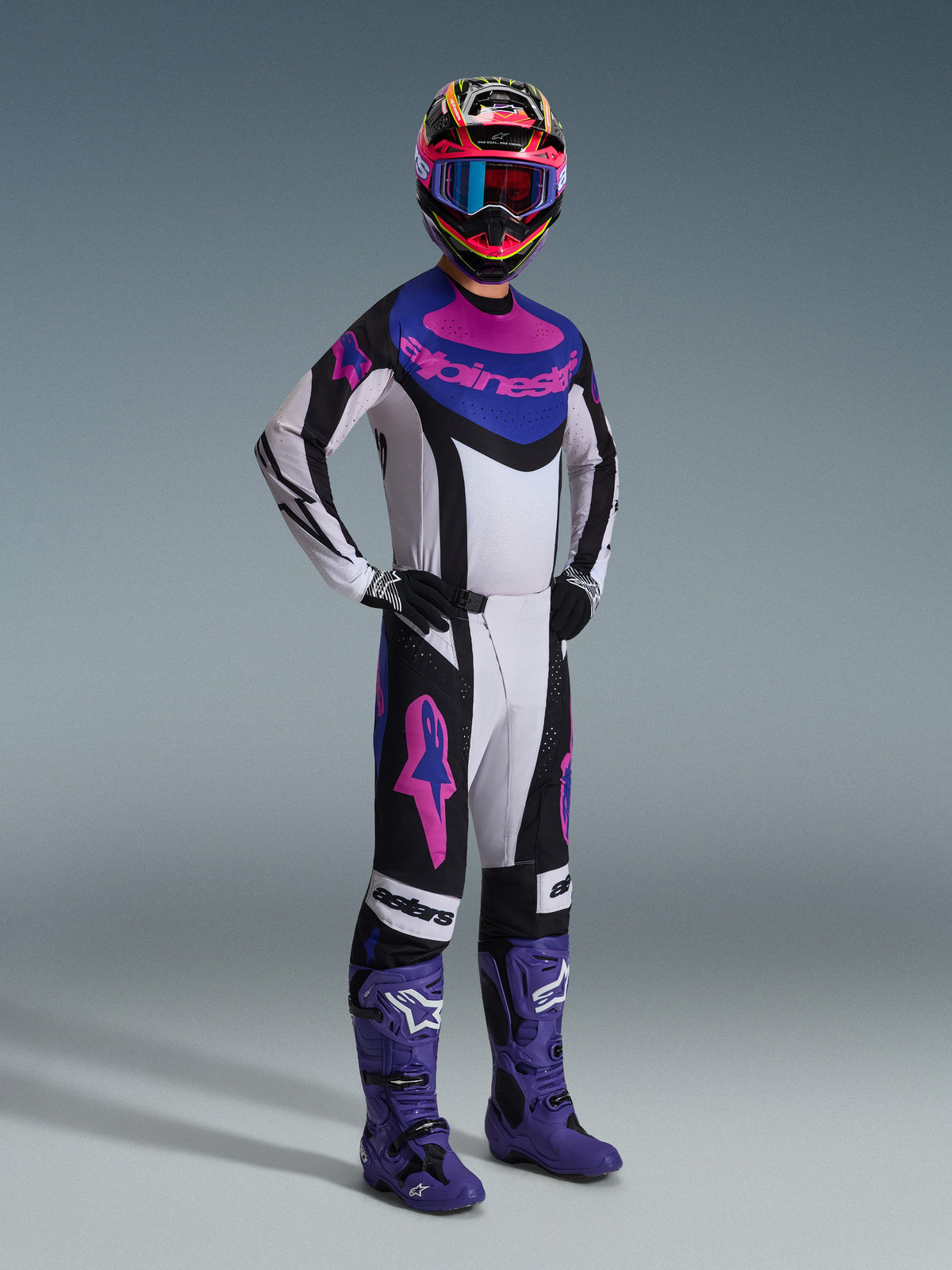 Pilota di motocross con il set completo Alpinestars Techstar Knif 2026, maglia bianca con dettagli neri, blu e rosa, pantaloni coordinati con loghi Astars rosa, casco multicolore con occhiali, stivali da gara viola, abbigliamento tecnico ad alte prestazioni per motocross