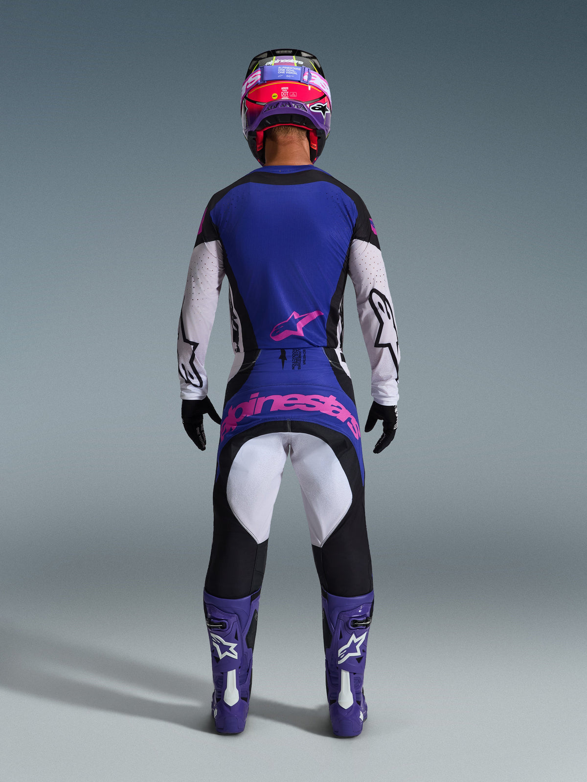 Vista posteriore di un pilota di motocross che indossa il set completo Alpinestars 2026 Techstar Knif, maglia blu e bianca con logo a stella rosa, pantaloni coordinati blu e neri con grande marchio Alpinestars rosa sul retro, stivali viola e bianchi e casco viola e rosso, abbigliamento professionale da fuoristrada coordinato