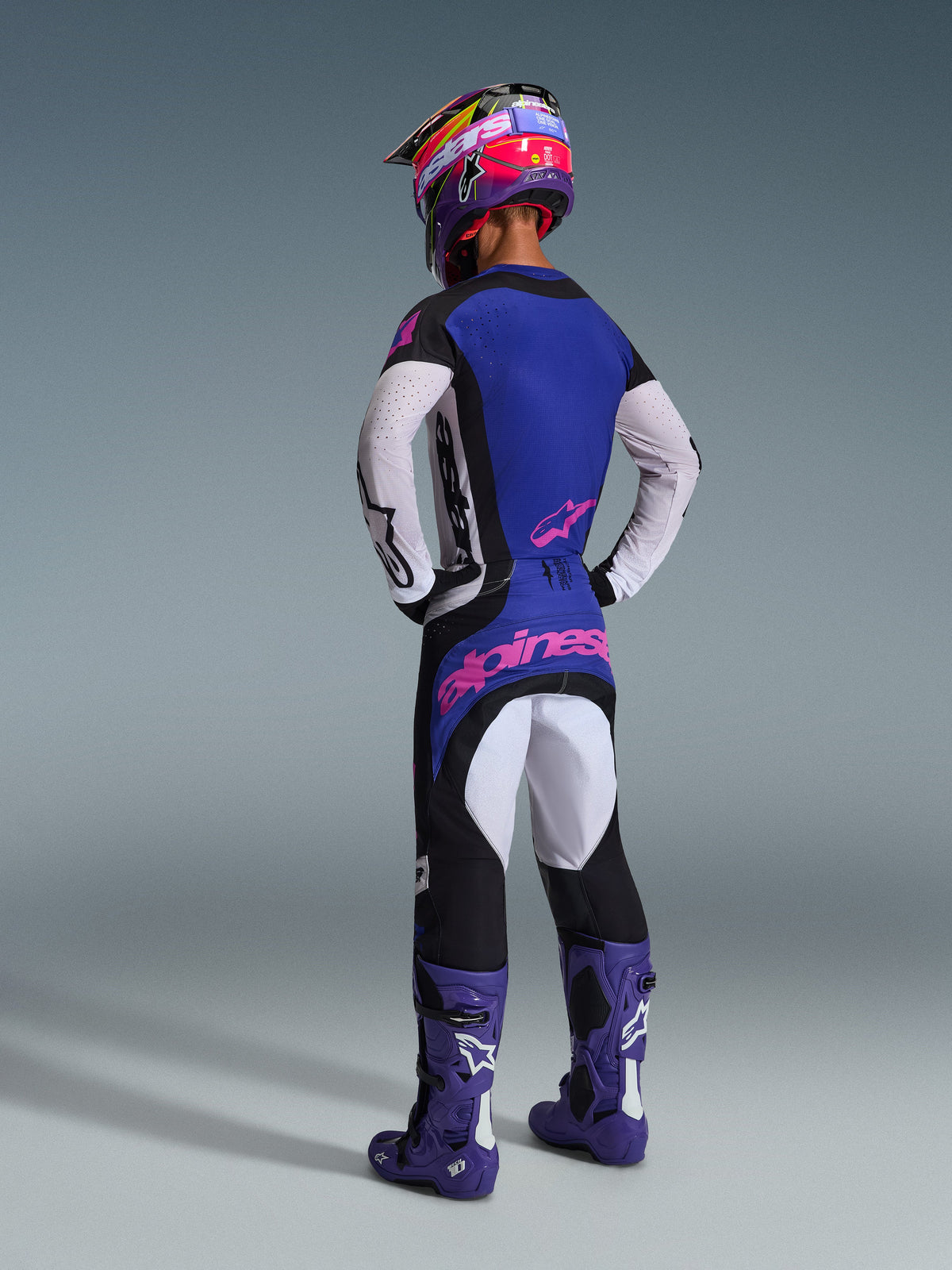 Pilota di motocross visto da dietro con il completo Alpinestars 2026 Techstar Knif, maglia blu con maniche bianche traforate e loghi rosa, pantaloni neri e bianchi con branding rosa sul retro, stivali viola e un vivace casco viola e rosa, outfit professionale da gara.