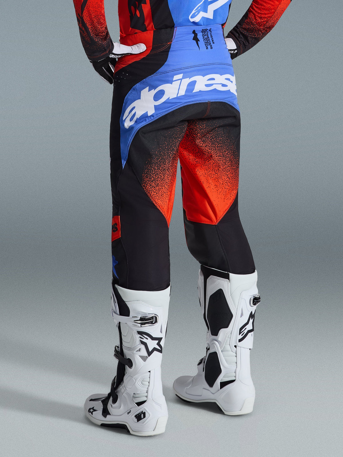 Alpinestars Pantaloni Techstar Knif 2026, pantaloni da motocross, arancione, nero e blu UCLA, vista posteriore con carré blu e logo Alpinestars bianco, motivo sfumato arancione sulle gambe, indossati con maglia coordinata e stivali bianchi