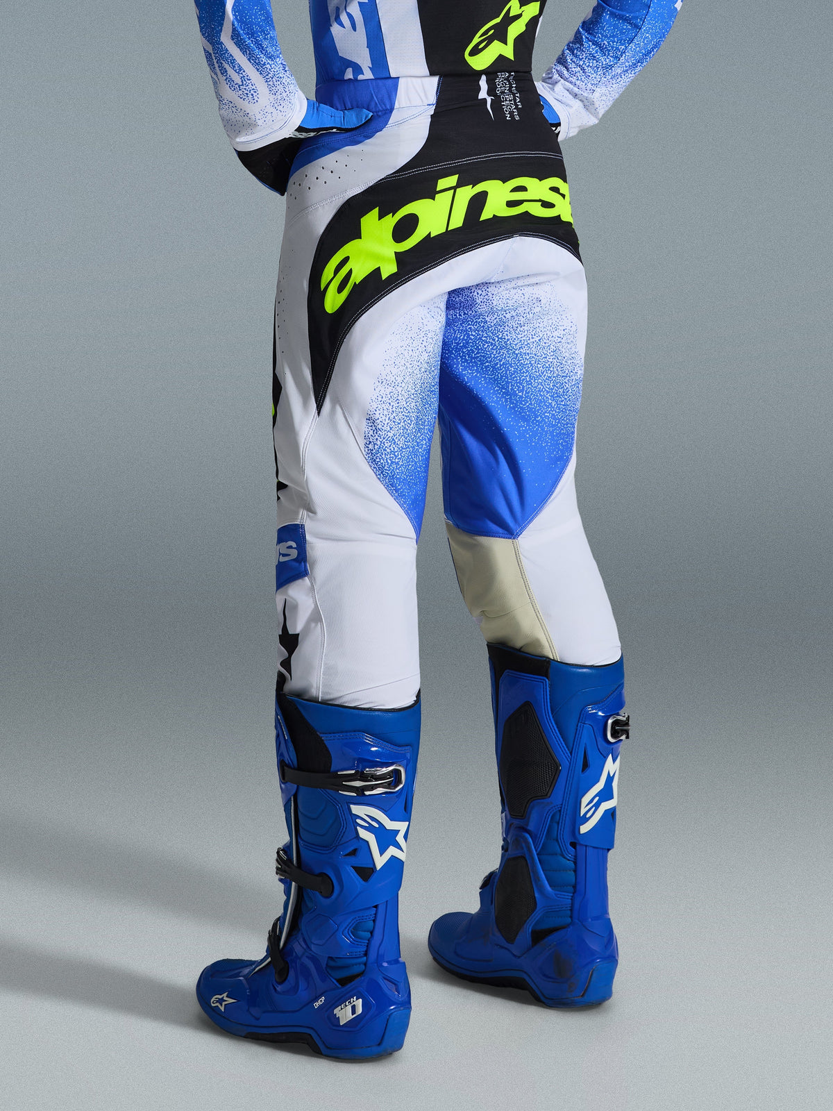 Alpinestars 2026 Techstar Knif Pants, pantaloni da motocross, blu UCLA, nero e giallo fluo, vista posteriore con pannelli bianchi e motivo sfumato blu a effetto spray, branding giallo fluo sulla seduta nera, indossati con stivali Tech 10 blu coordinati e maglia per il fuoristrada.