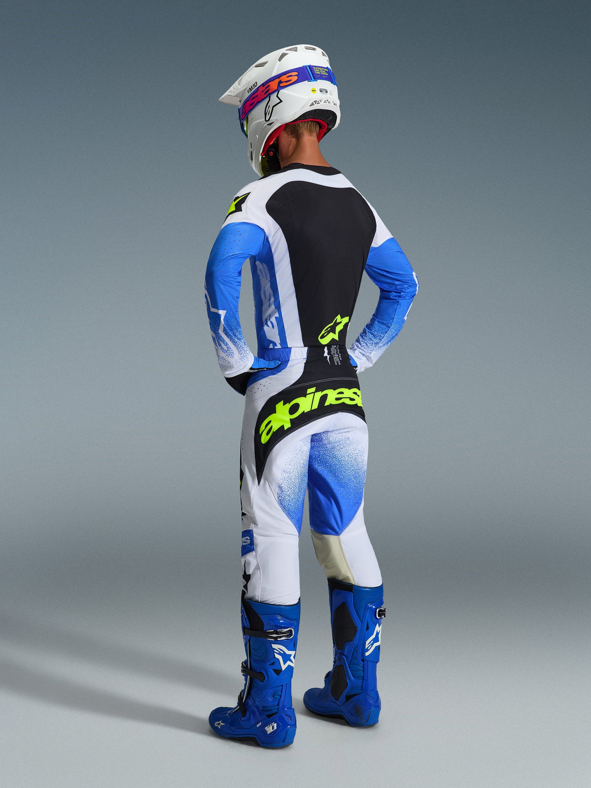 Pilota di motocross inquadrato da dietro che indossa i pantaloni Techstar Knif 2026 in Ucla Blue Black e Yellow Fluo, con logo giallo acceso sulla seduta, maglia coordinata con maniche blu, casco bianco e stivali da fuoristrada blu.