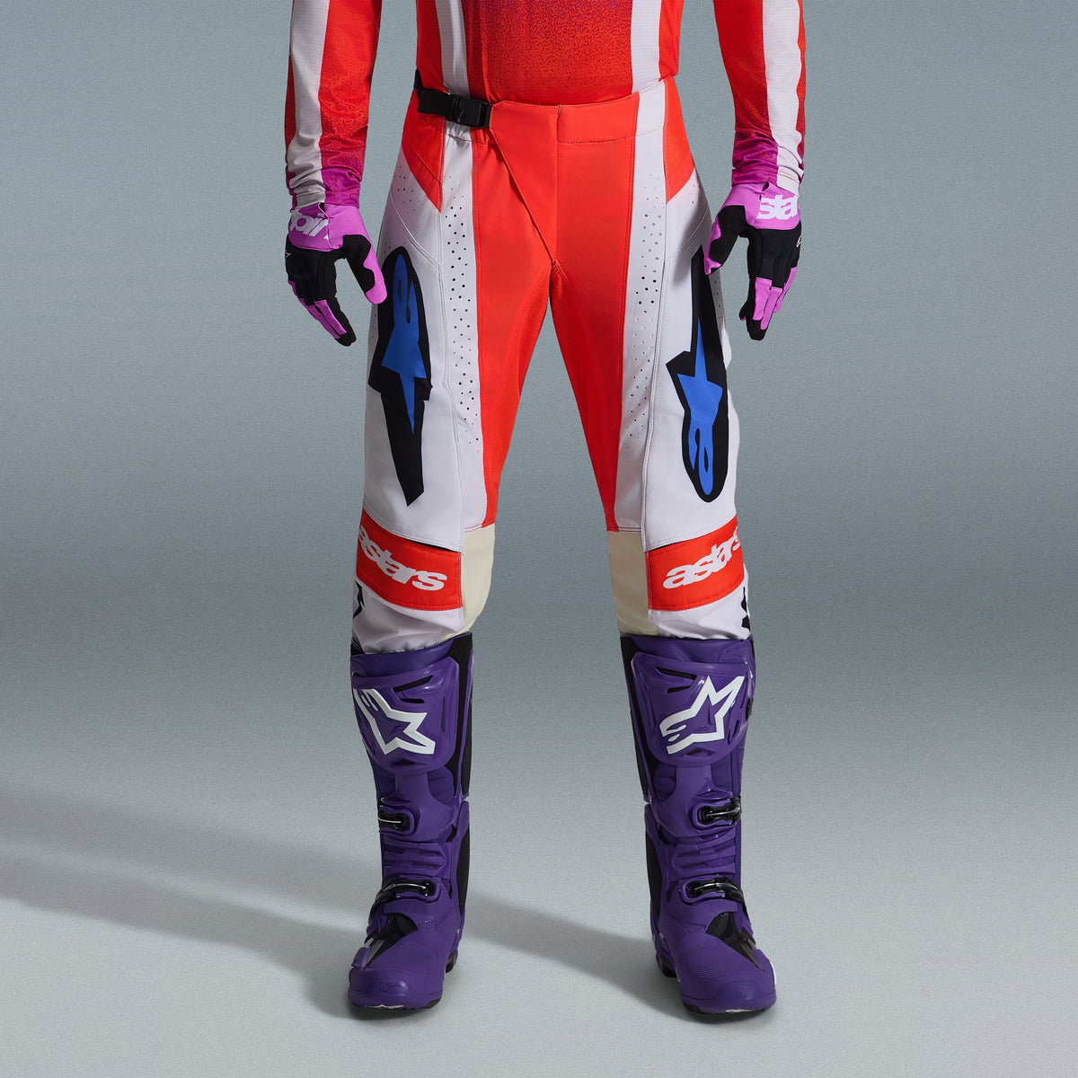Alpinestars 2026 Techstar Knif Gear Set, parte inferiore del corpo di un pilota di motocross che indossa pantaloni rossi e bianchi con accenti blu, stivali da gara viola acceso con loghi bianchi e guanti coordinati neri e viola, kit professionale da gara per fuoristrada